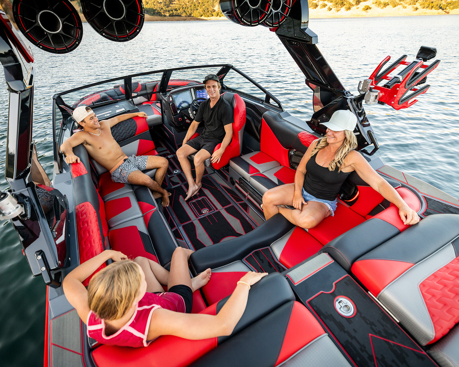 Four people sit and relax on a red and black Centurion Ri230, a top-tier wake boat, on a lake, with water sports equipment attached to the boat.