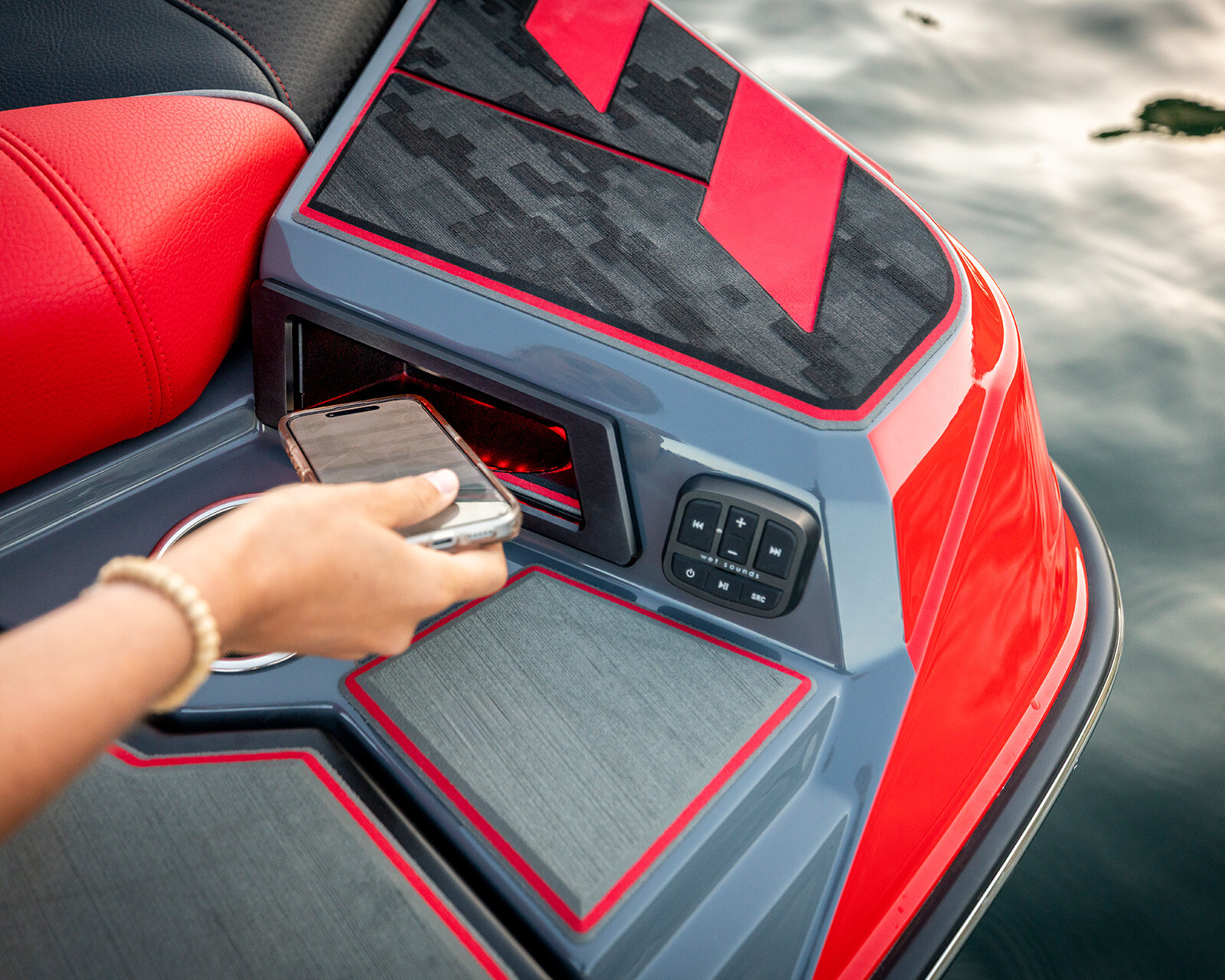A person places a smartphone into a storage compartment on a red and gray Centurion Ri230 wake boat, with control buttons and water visible nearby.