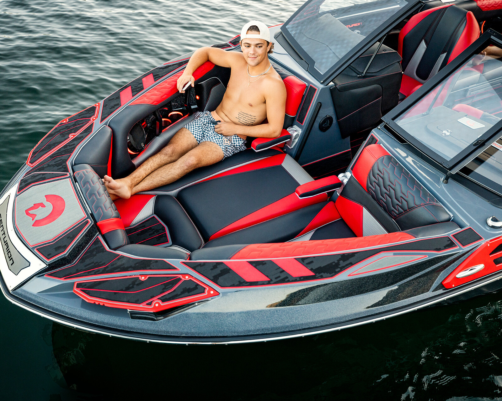 A shirtless man in patterned shorts and a white cap sits relaxed on a sleek, red and black Centurion Ri230 speedboat floating on the water.
