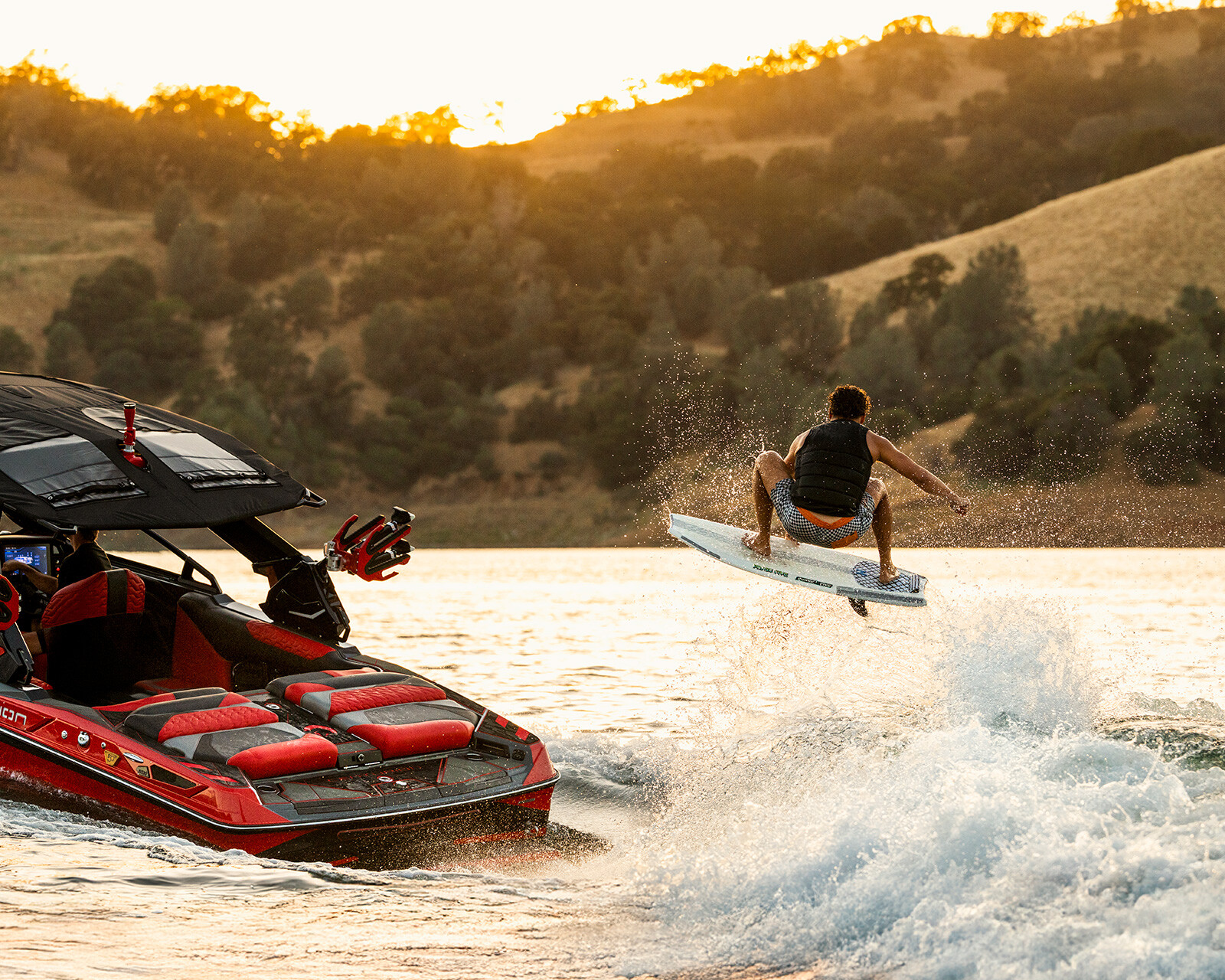 A person in a life vest rides a wakeboard, soaring over the water behind a Centurion Ri230 as it speeds across the lake at sunset, framed by scenic hills and trees.
