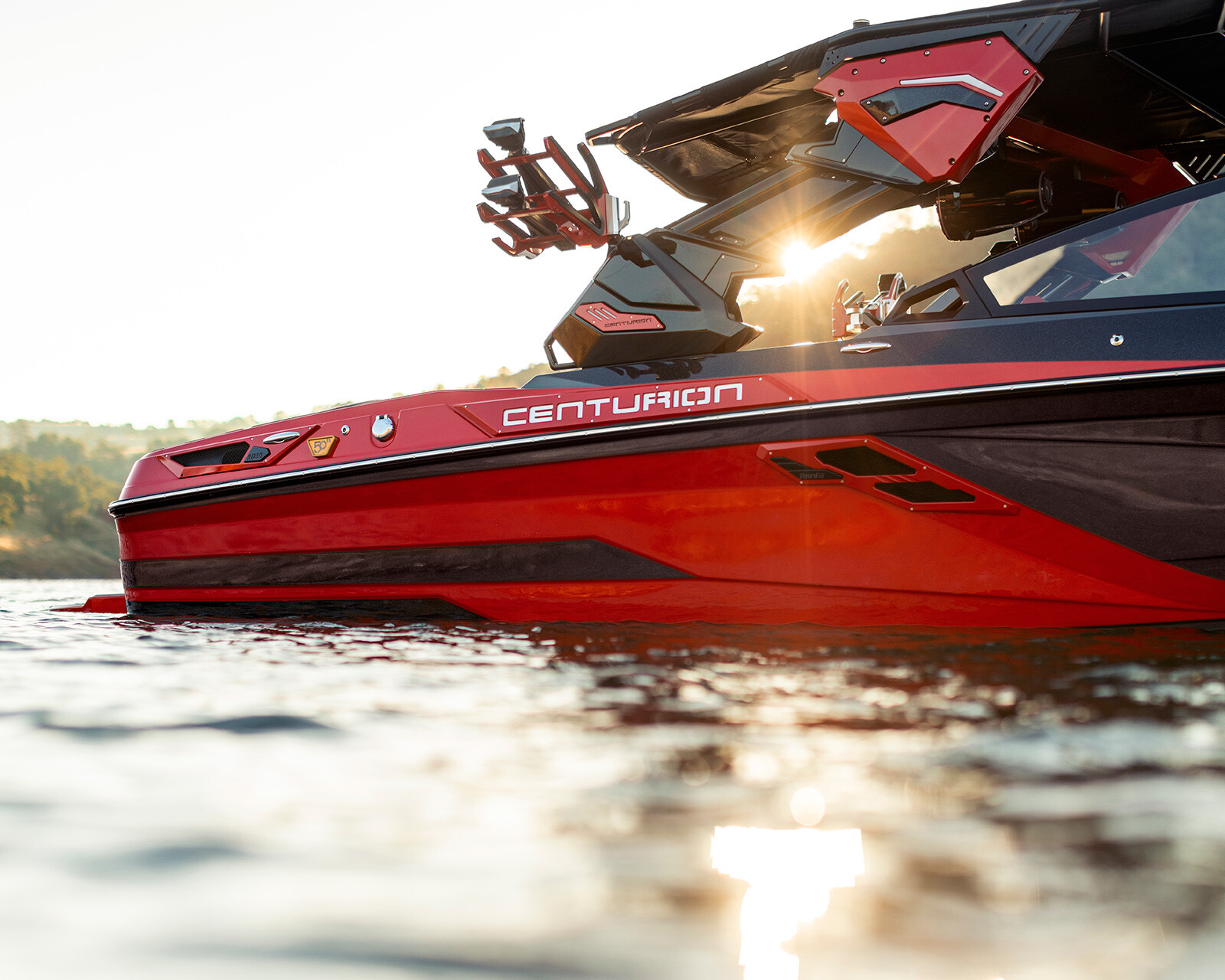 A red Centurion Ri230 Wake Boat floats on calm water at sunset, with sunlight shining through its open canopy.