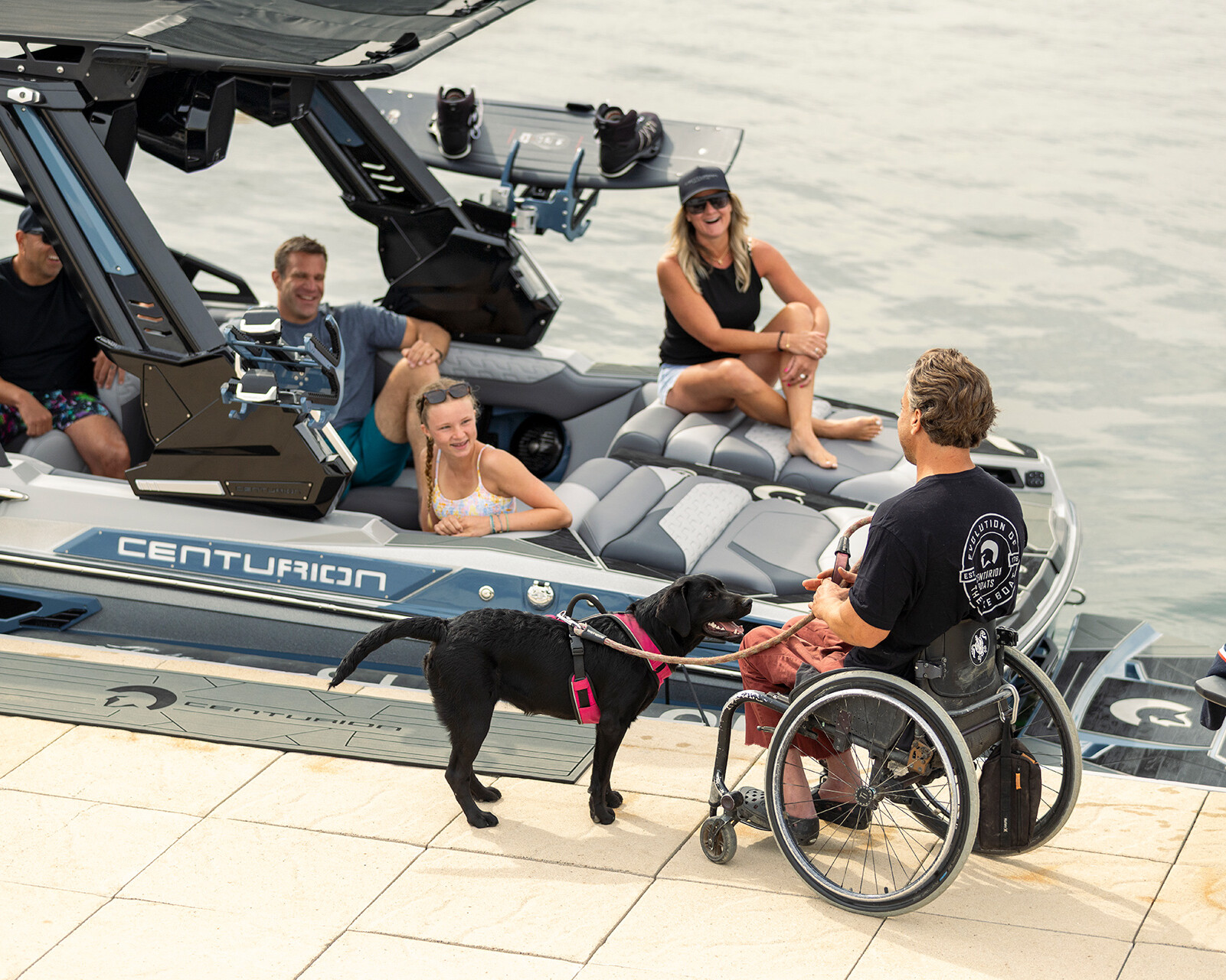 A person in a wheelchair with a service dog talks to four people on a boat docked at the water's edge beside a sleek Centurion Ri230.