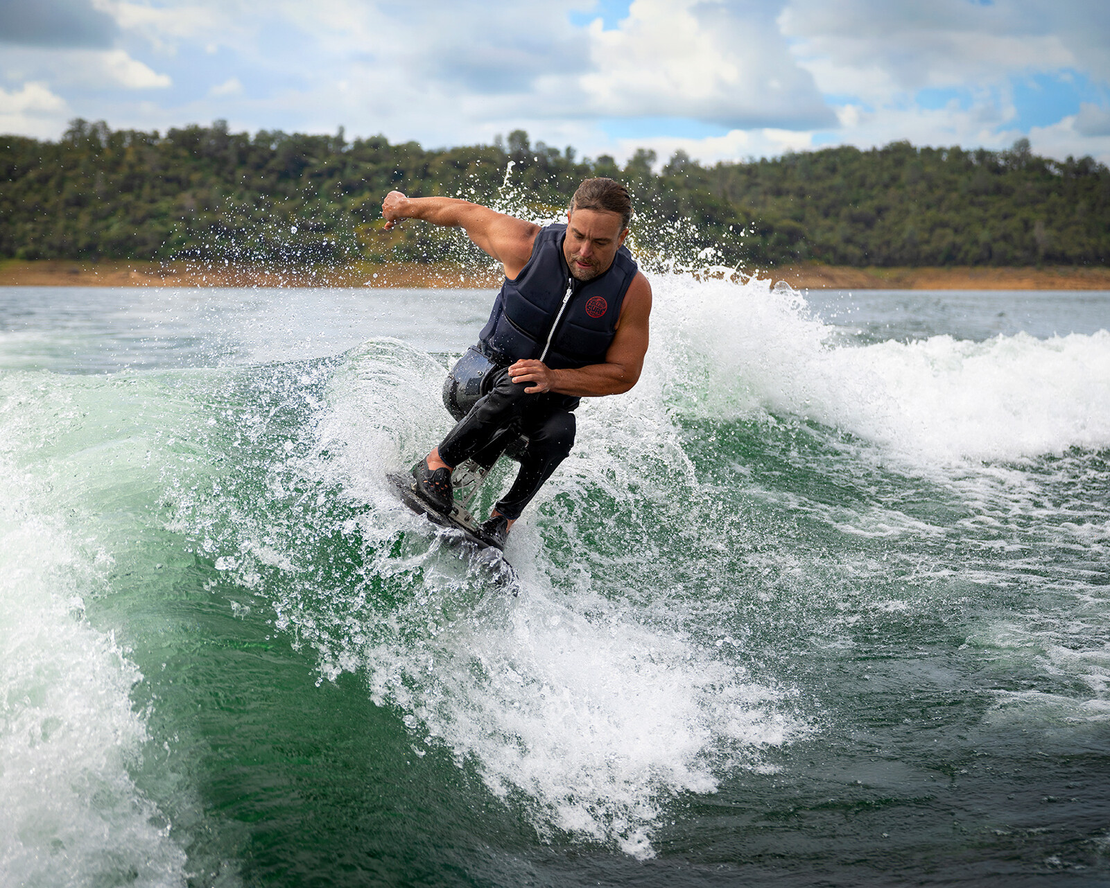 A man in a wetsuit rides a wave behind a Centurion Ri230 surf boat, with a forested shoreline and cloudy sky in the background.