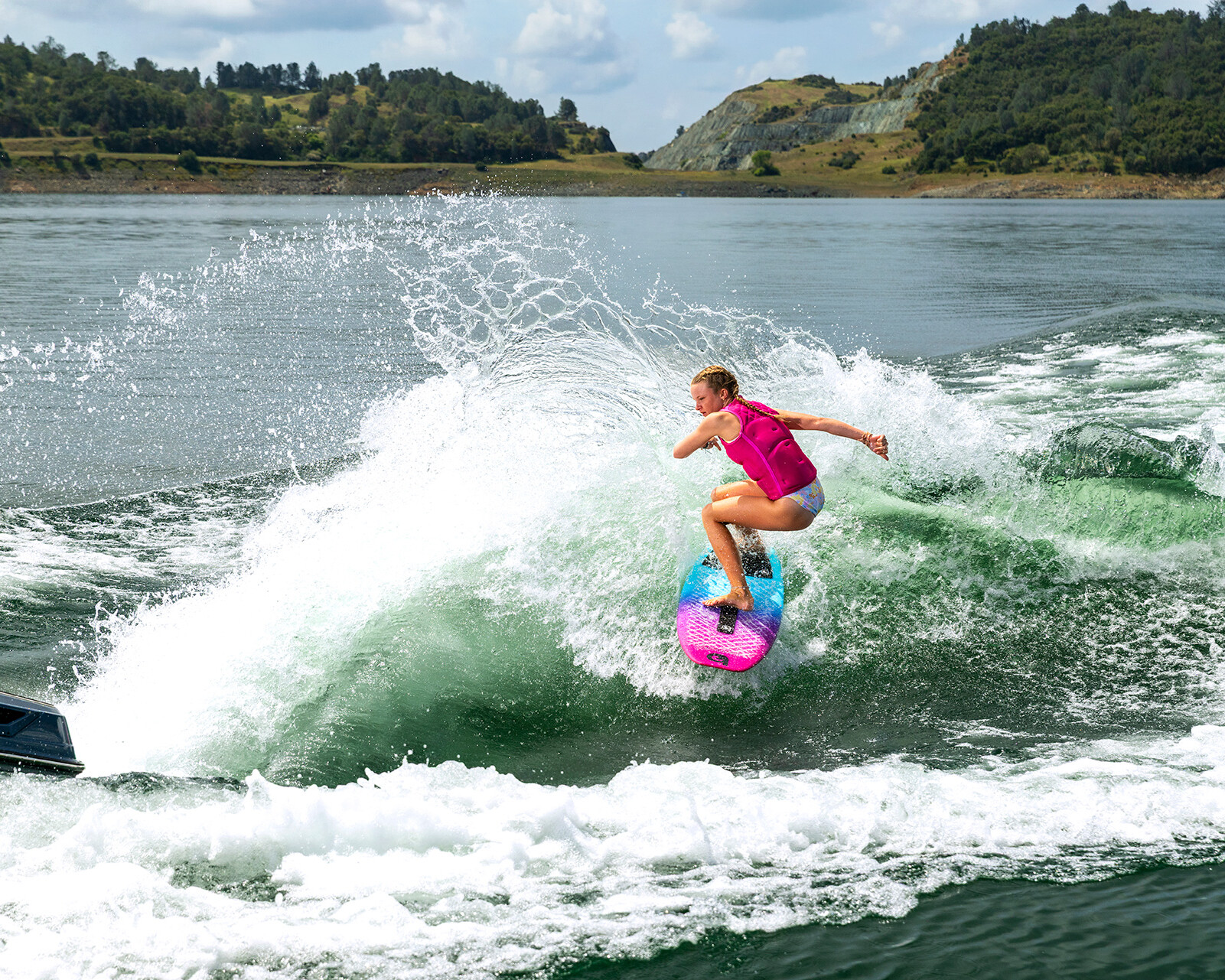 A person in a pink vest rides a wave on a blue and pink surfboard behind a Centurion Ri230 Wake Boat on a lake, with green hills in the background.