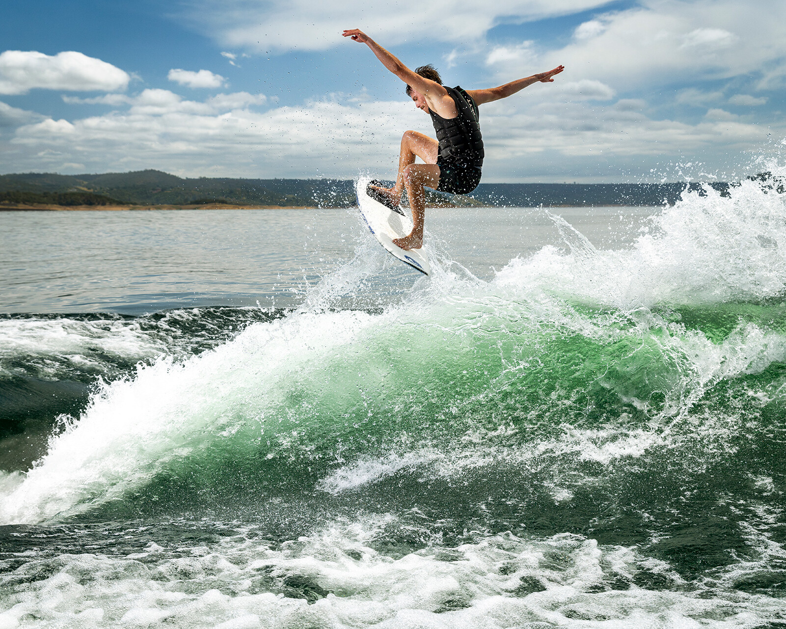 A person in a black outfit is performing a jump on a surfboard over a wave created by a Centurion Ri230, with water splashing under a partly cloudy sky.