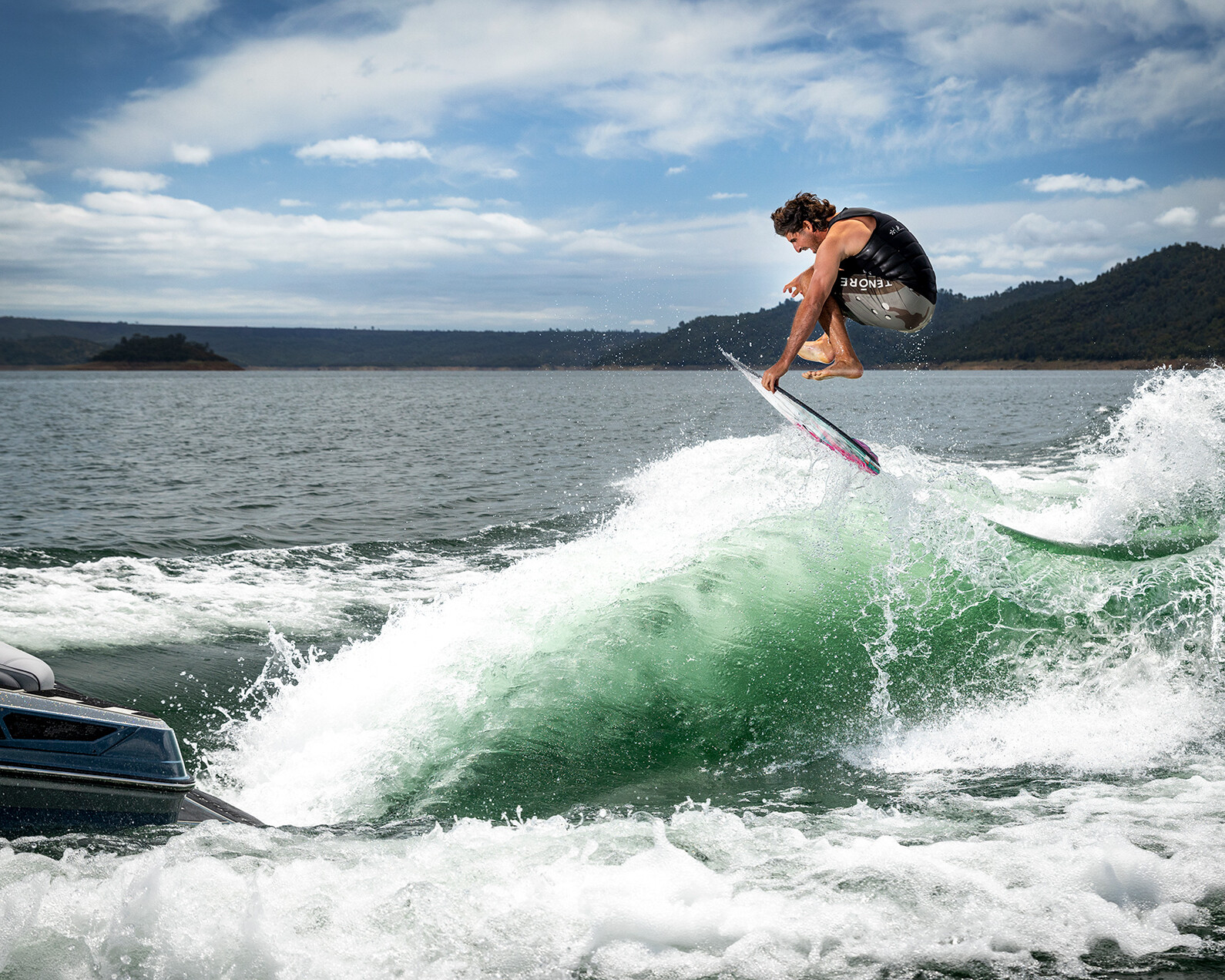 A man in a life vest performs a jump on a wakesurf board behind a Centurion Ri230, carving through green waves with hills in the background.
