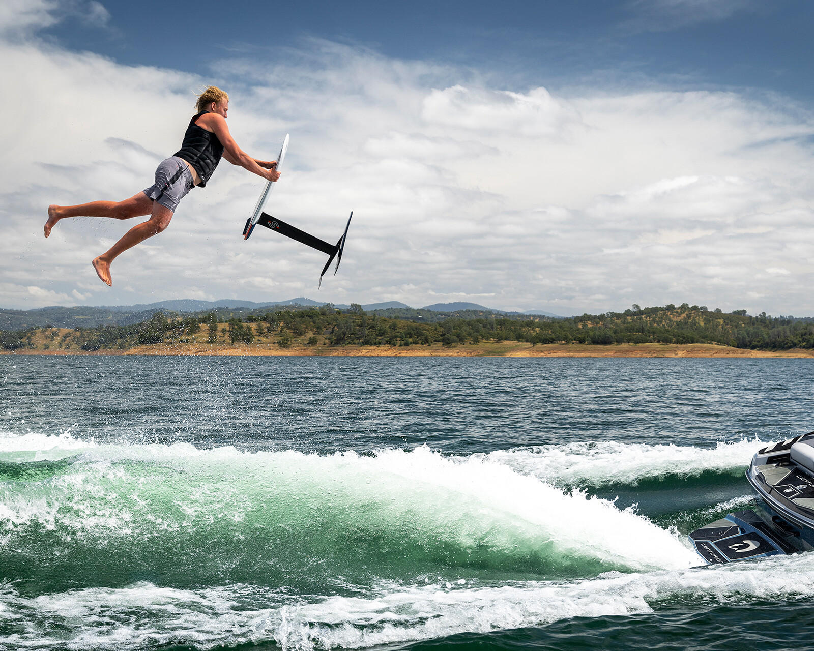 A person is airborne above the water, holding a hydrofoil board, after hitting a wave created by a Centurion Ri230 Surf Boat.