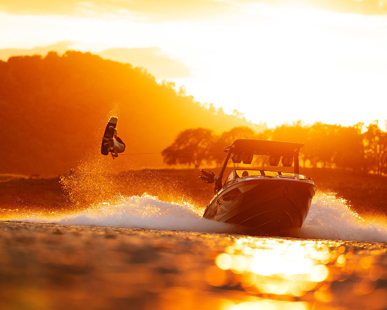 A person wakeboards behind a Centurion Ri230 on a lake at sunset, with trees and hills silhouetted in the background.