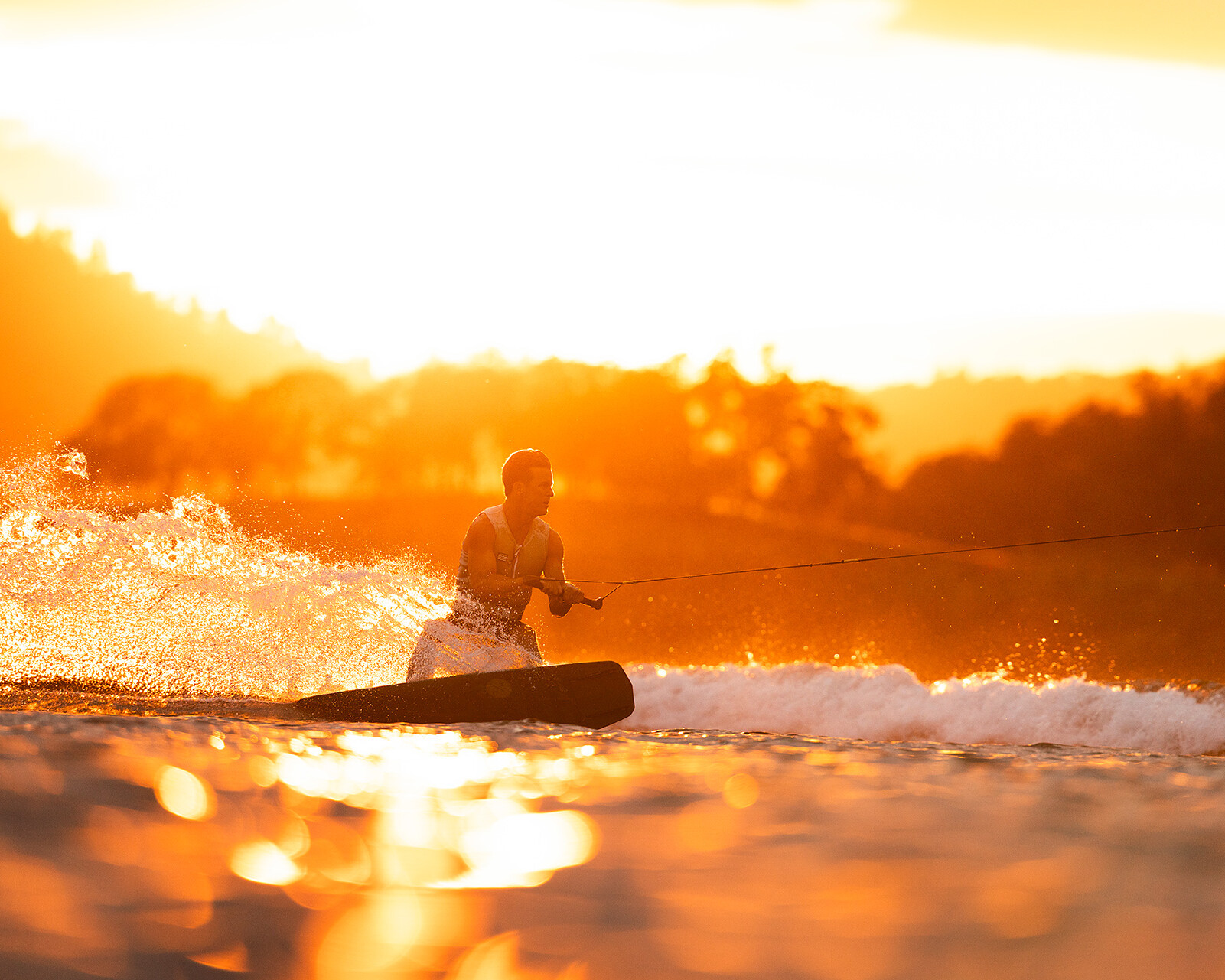 A person wakeboarding on a lake at sunset behind a Centurion Ri230, creating splashes with golden light reflecting on the water.