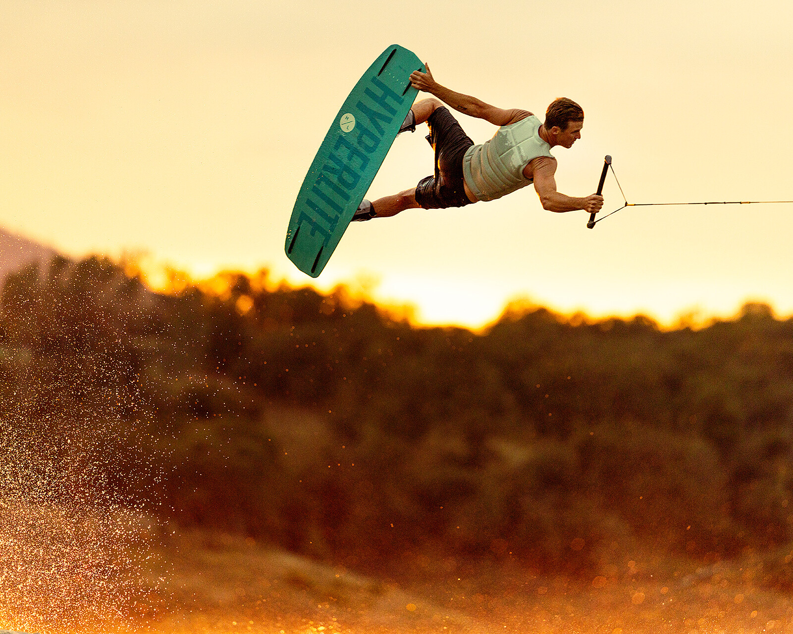 A person wakeboarding behind a Centurion Ri230 performs an aerial trick while holding a handle, with water spray and trees visible in the background at sunset.