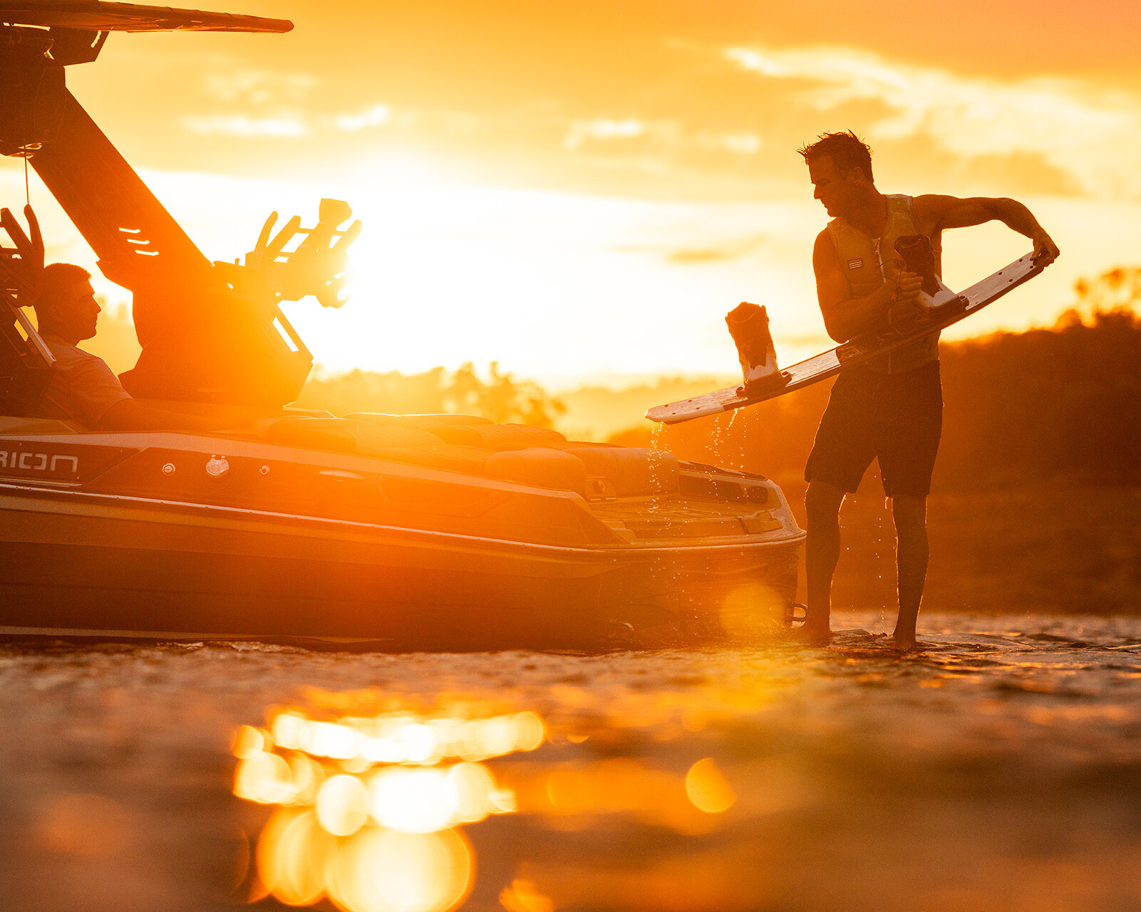 A person holding a wakeboard stands in shallow water next to a Centurion Ri230, with sunlight reflecting off the water's surface at sunset.