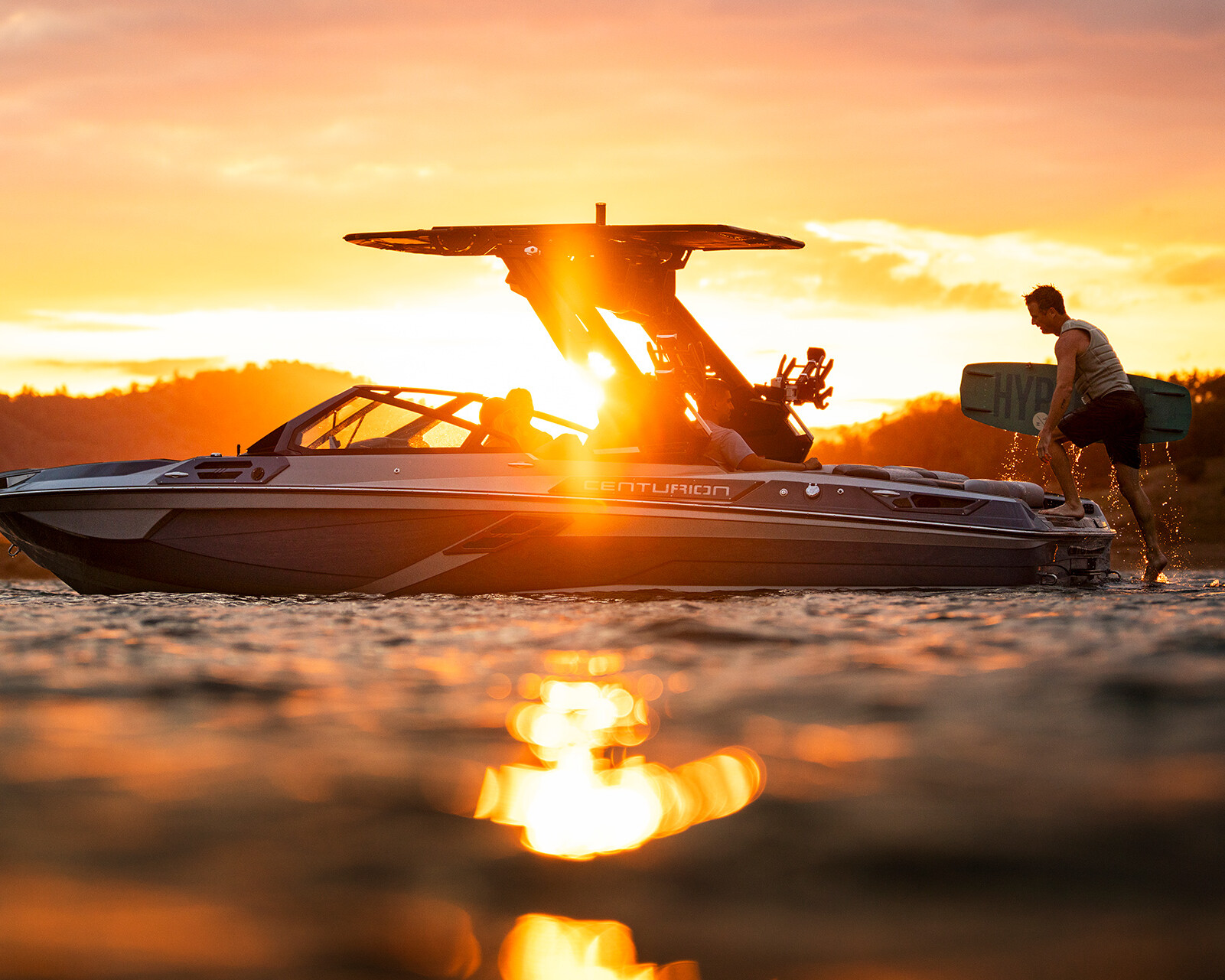 A person stands on the rear platform of a Centurion Ri230 at sunset, preparing a wakeboard as sunlight reflects off the water.