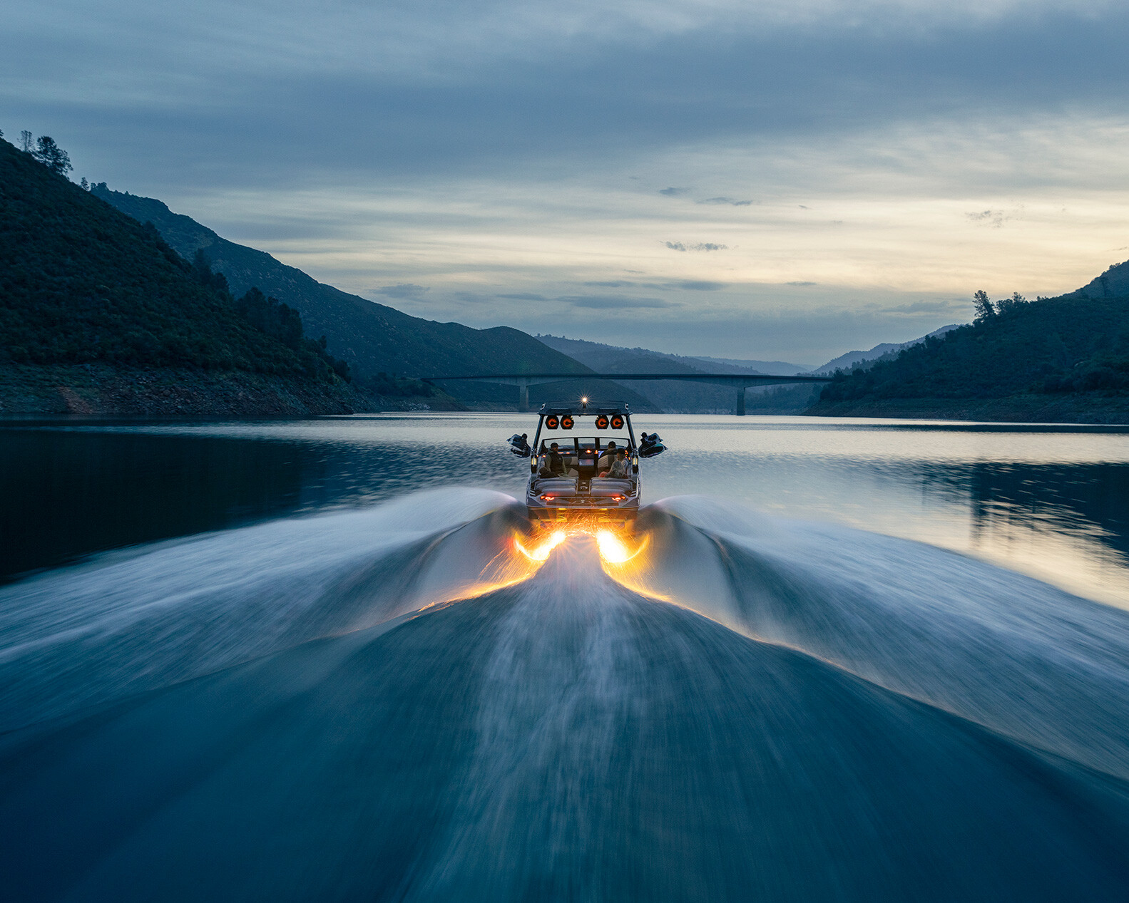 A Centurion Ri230 glides quickly across a calm lake at dusk, leaving a bright, illuminated wake behind it, with the silhouette of hills and a bridge in the background.
