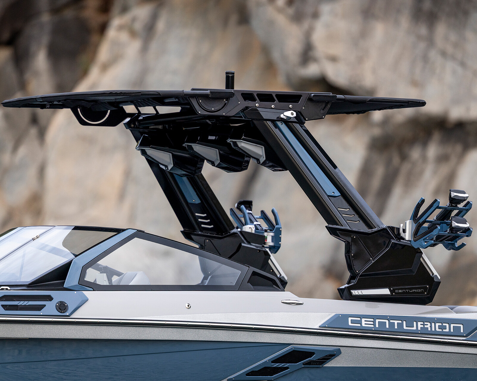 A close-up view of a Centurion Ri230 Surf Boat featuring a sleek black wakeboard tower with racks, set against a rocky background.