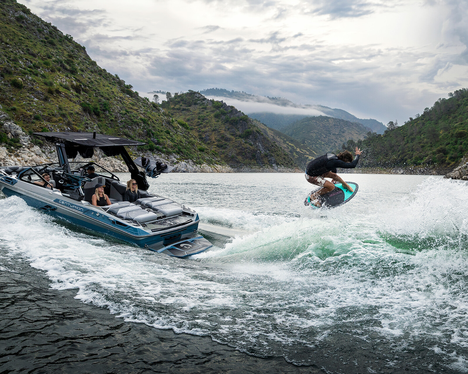 A person wakesurfs behind a Centurion Ri230 on a lake surrounded by hills, while three people watch from the boat under a cloudy sky.