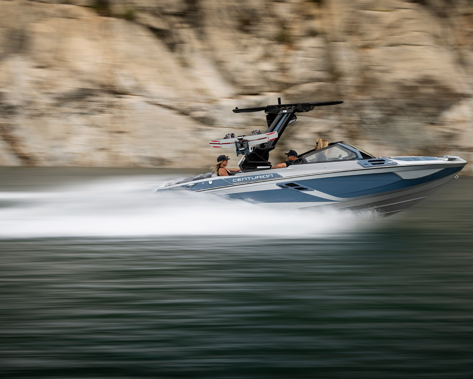 A blue and white Centurion Ri230 Wake Boat moves quickly across the water near a rocky shoreline, leaving a blurred wake behind it.