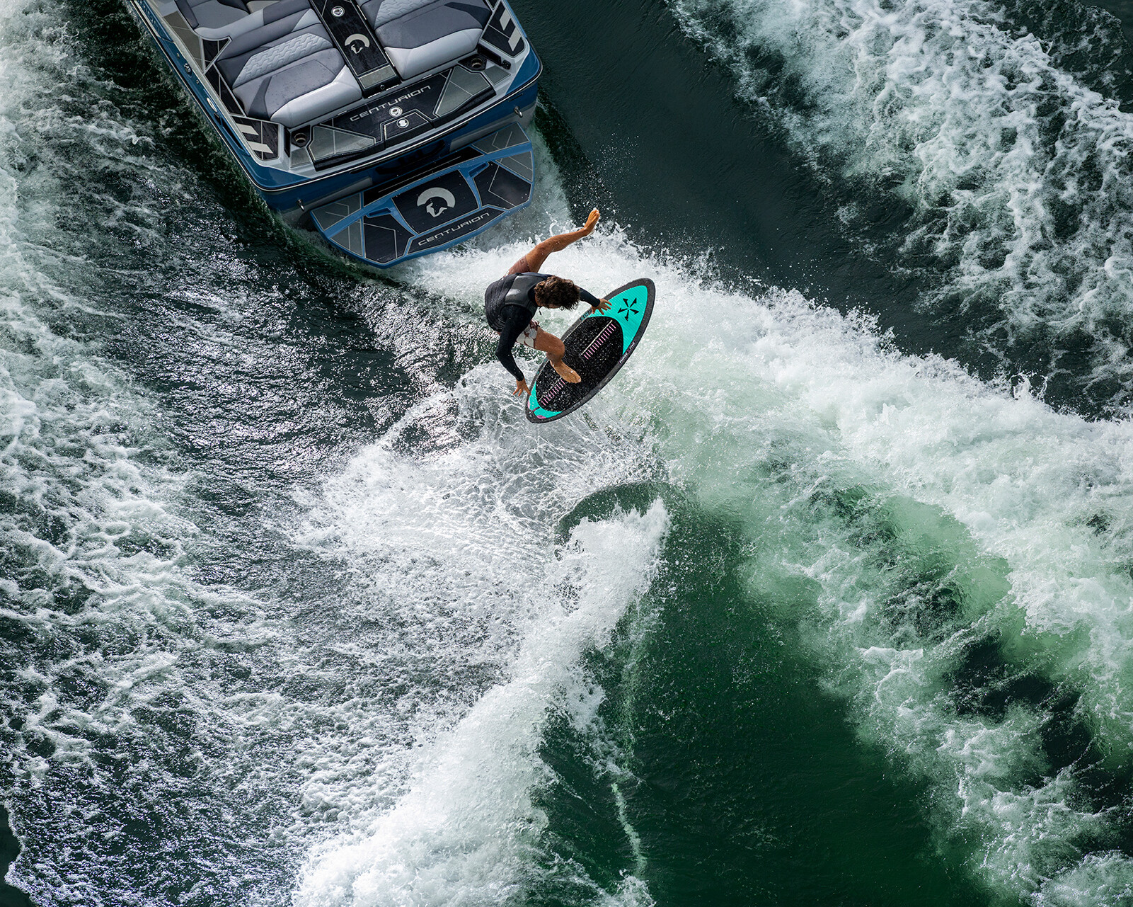 A person on a wakesurf board rides the wave behind a Centurion Ri230 wake boat on a green body of water, captured from above.