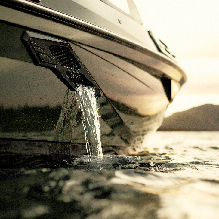 Close-up view of water flowing out of a drainage outlet on the side of a boat, with the boat partially submerged and a calm, sunlit lake in the background.