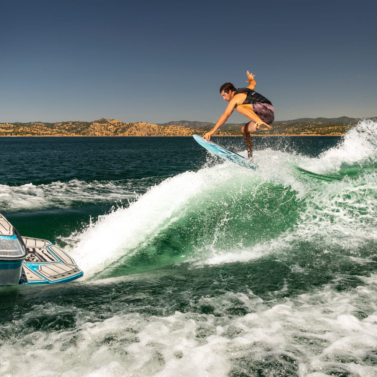 A man in a life vest is wakesurfing on a wave created by a Centurion Ri230, gliding across the lake, surrounded by hills under a clear sky.