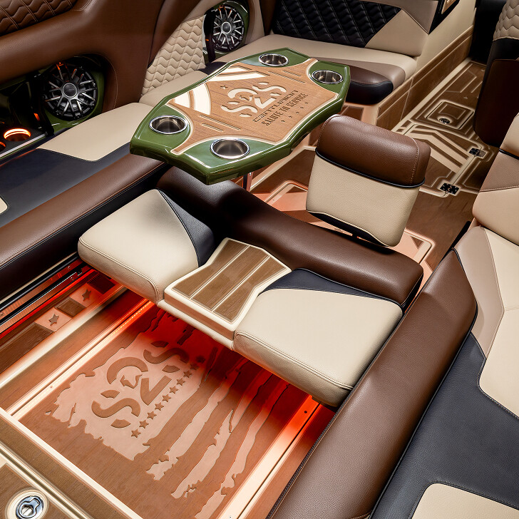 Luxury vehicle interior featuring custom leather seats, a wood and leather table with cup holders, and detailed flooring with an American flag design, all in brown, beige, and black tones.