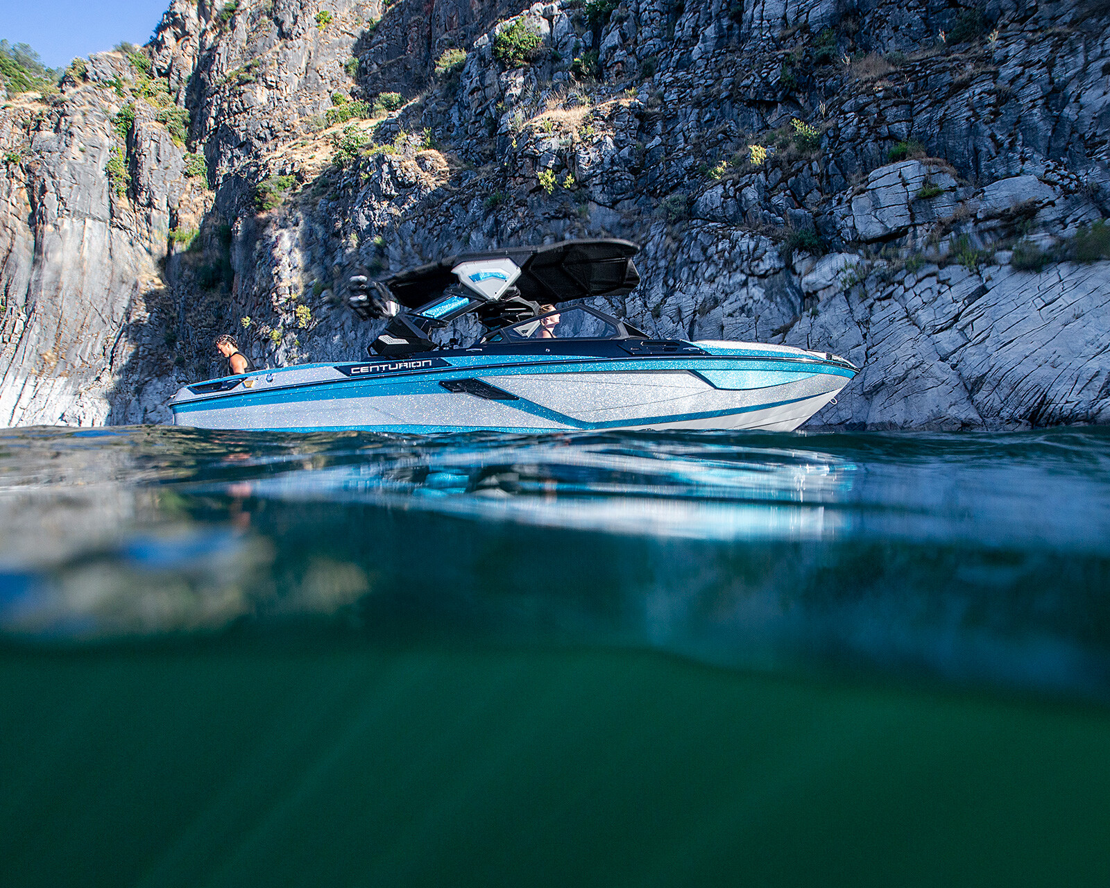 A blue and white Centurion Ri245 floats on clear water near rocky cliffs, with a person visible on board under a partly sunny sky.