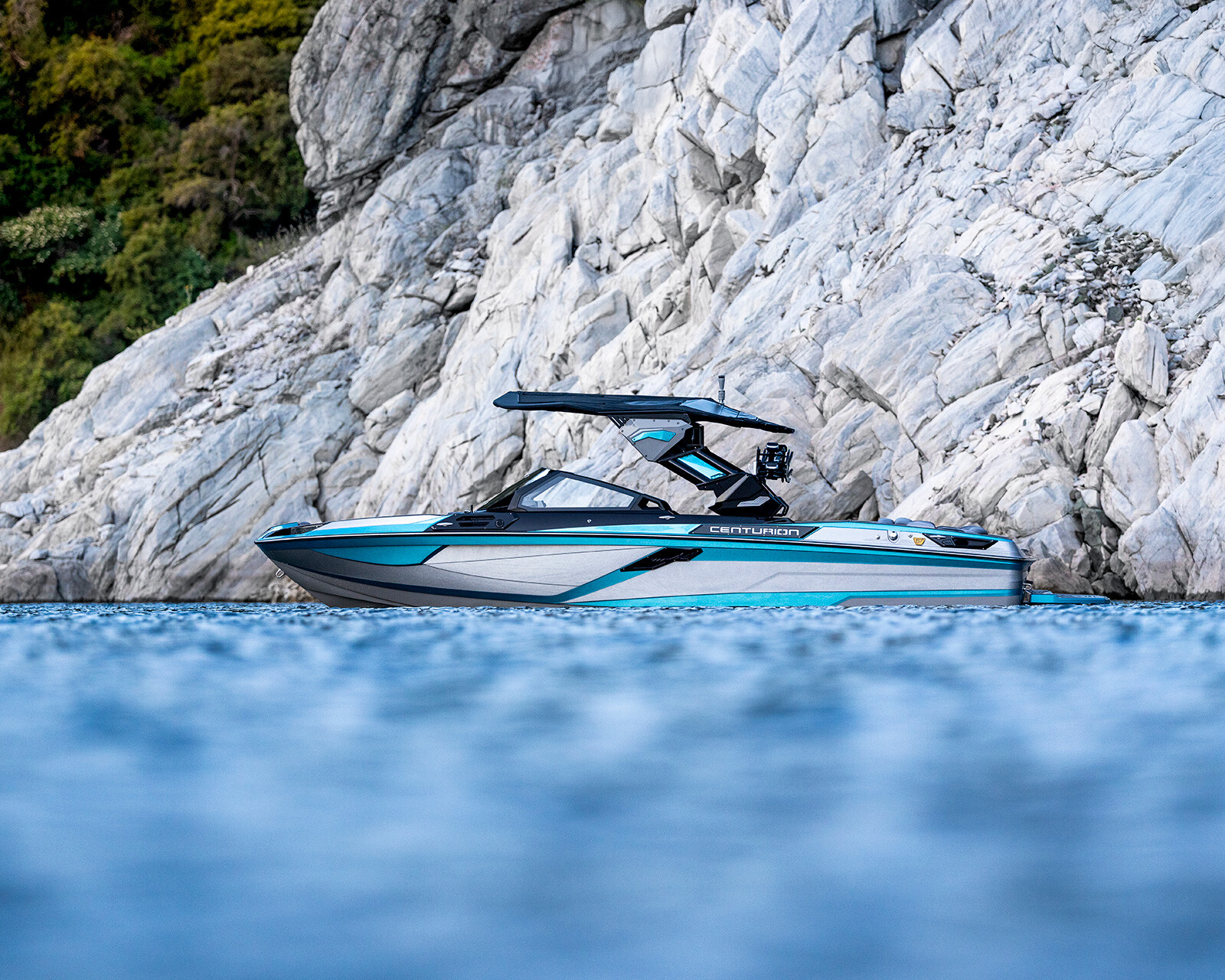 A modern Ri230 Wake Boat with blue and gray accents floats on calm water near a rocky white cliff adorned with greenery.