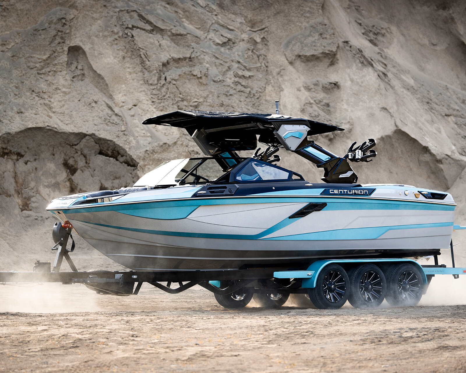 A white and blue Centurion Ri245 Surf Boat on a trailer hitched to a vehicle, parked on dusty ground near rocky cliffs.