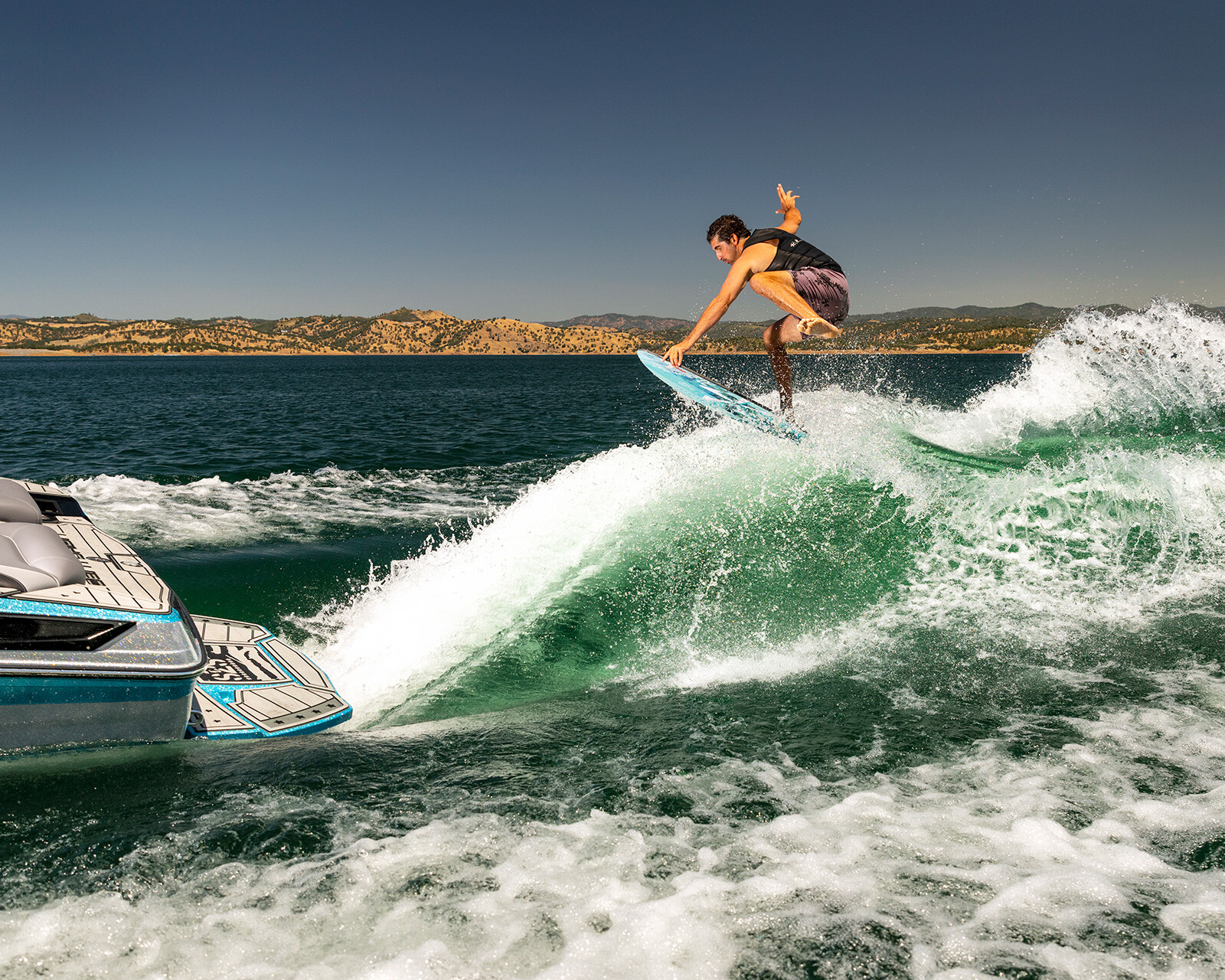 A person is wake surfing on a wave created by a Centurion Ri245 on a sunny day, with water splashing and hills visible in the background.