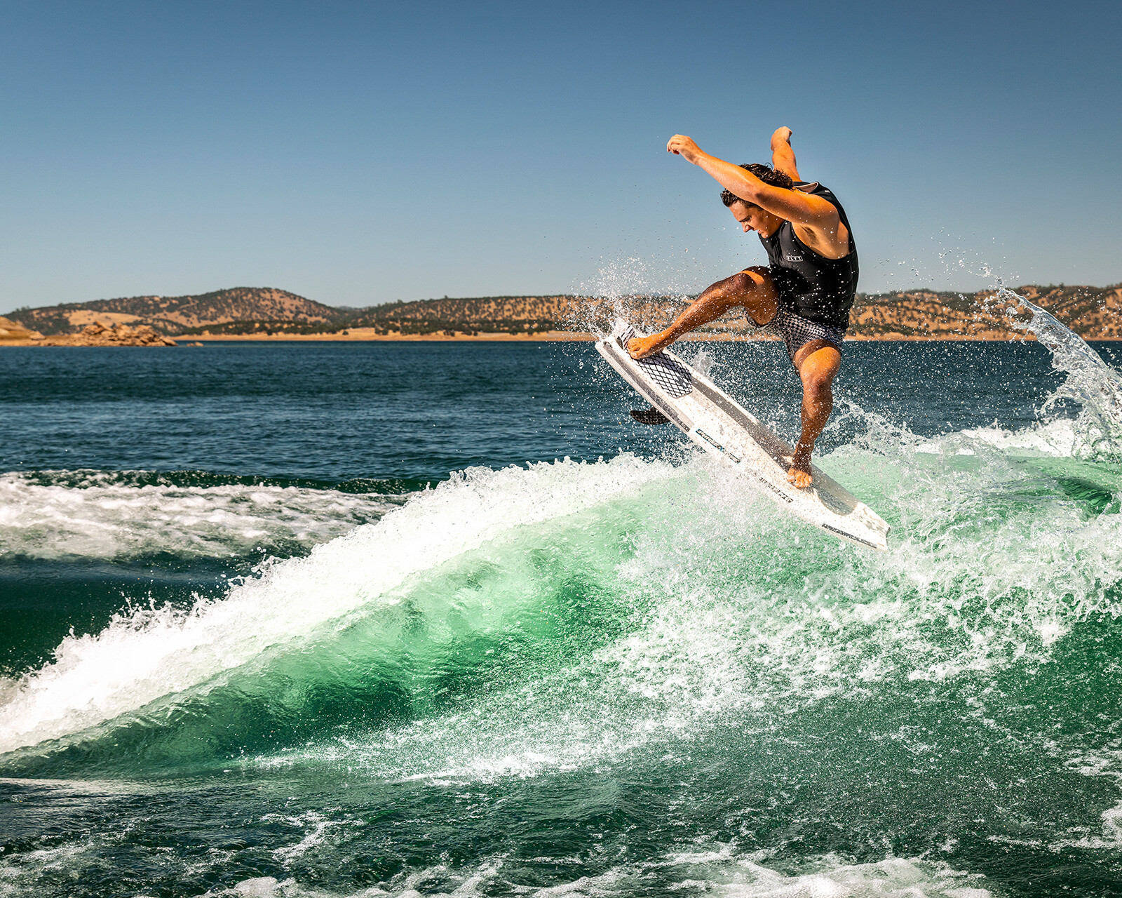 A person wearing a black vest performs an aerial trick on a wakeboard behind a Centurion Ri245 in a large body of water, with hills and clear blue sky in the background.