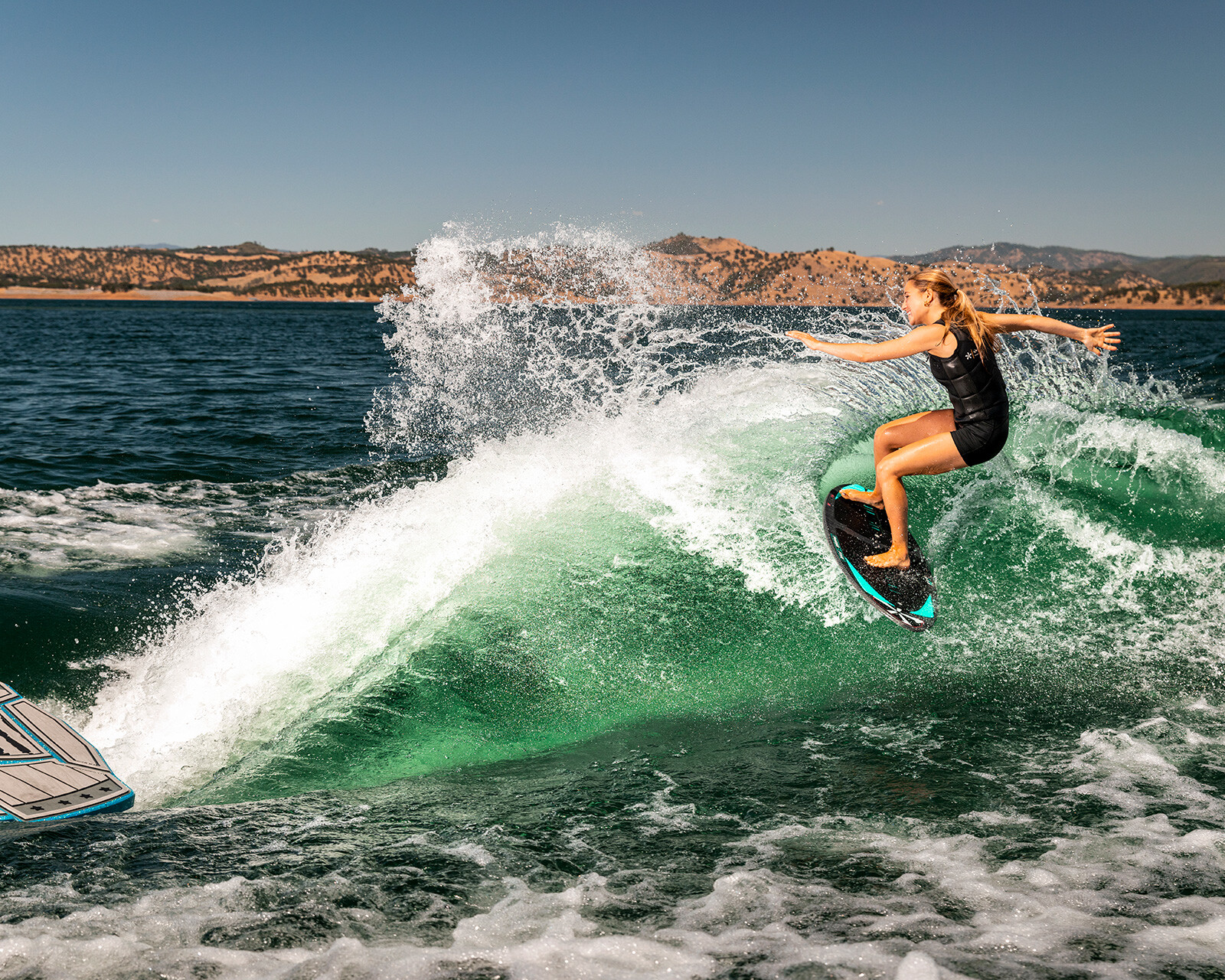 A woman in a black wetsuit is wakesurfing on a lake, riding a wave near a Centurion Ri245 surf boat, with mountains visible in the background.