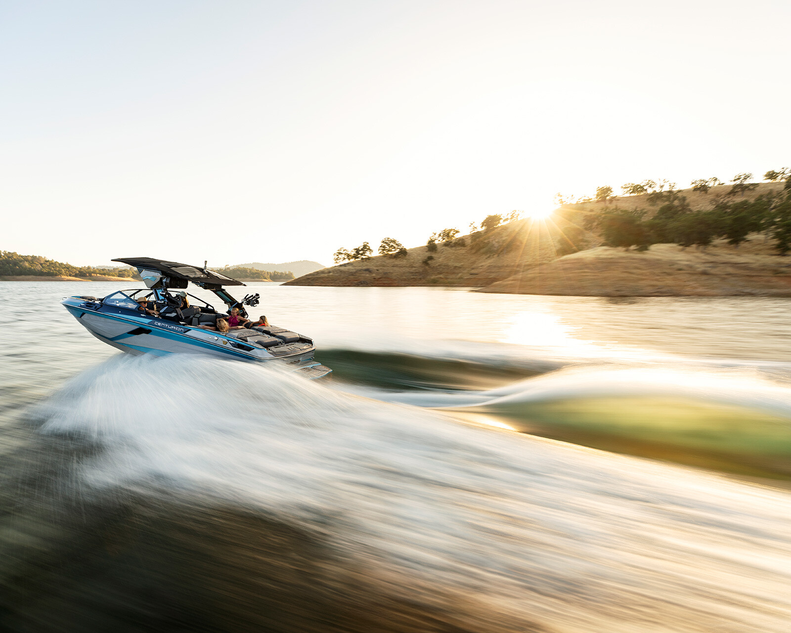A Centurion Ri245 glides swiftly across a lake near a sunlit shoreline, framed by trees and rolling hills in the background.