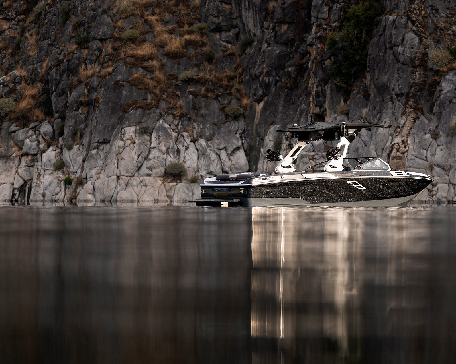 A black and white Centurion Ri265 wakeboard boat is stationary on calm water near a rocky cliff with sparse vegetation.