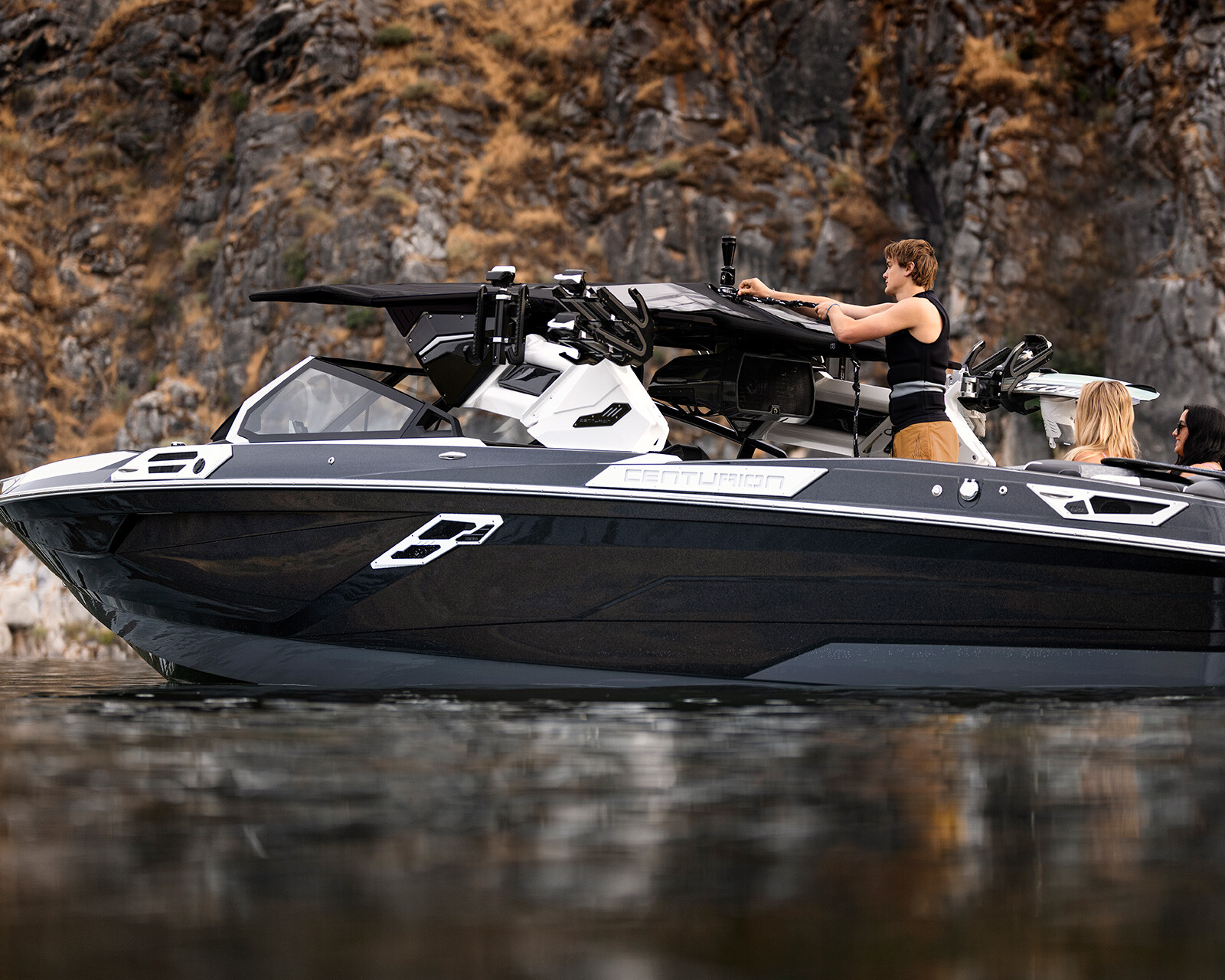 A group of people rides on a sleek black and white Centurion Ri265 Surf Boat near a rocky shoreline.