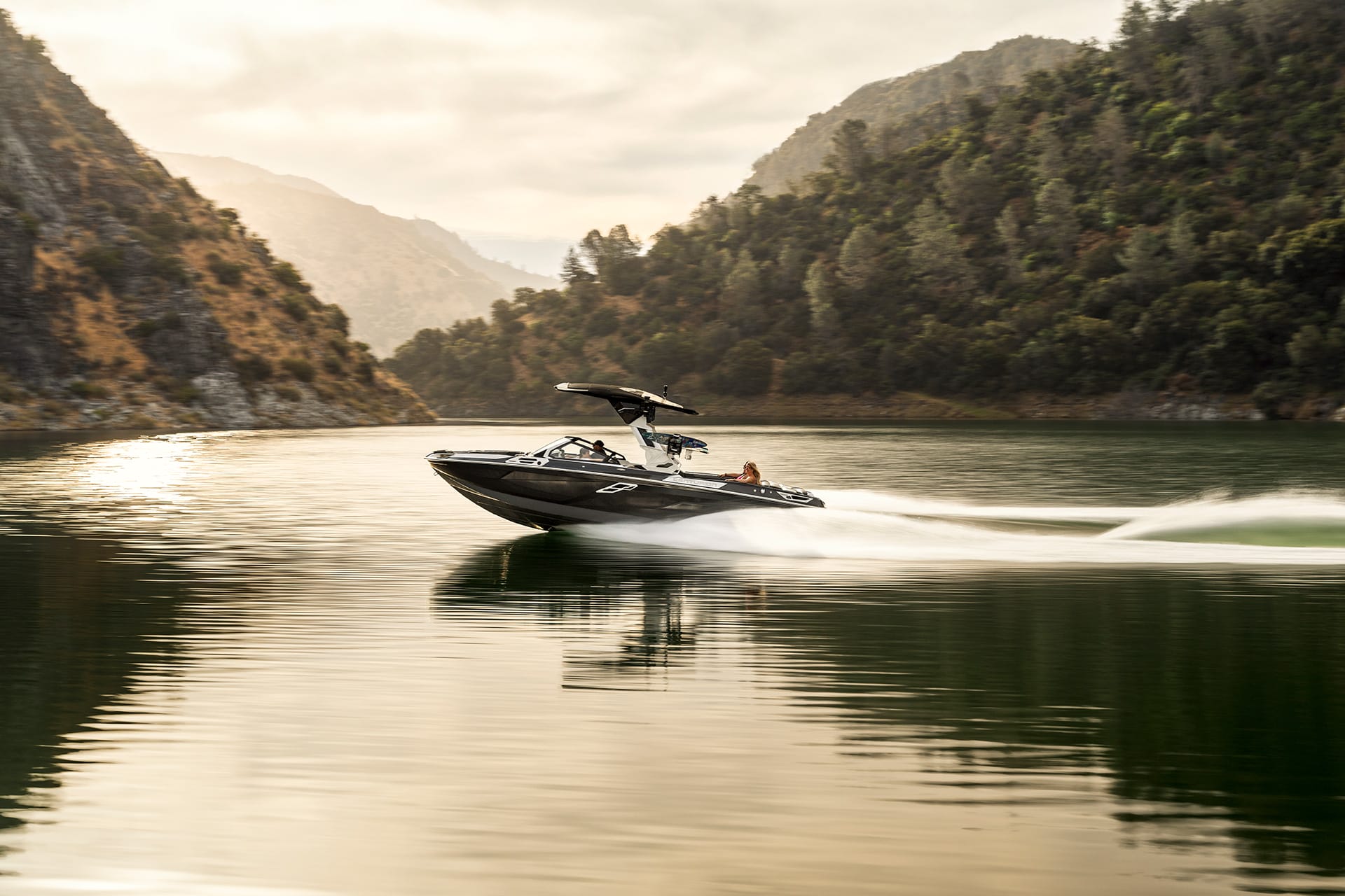 A black Centurion Ri265 glides quickly across a calm lake surrounded by forested hills and mountains under a cloudy sky.