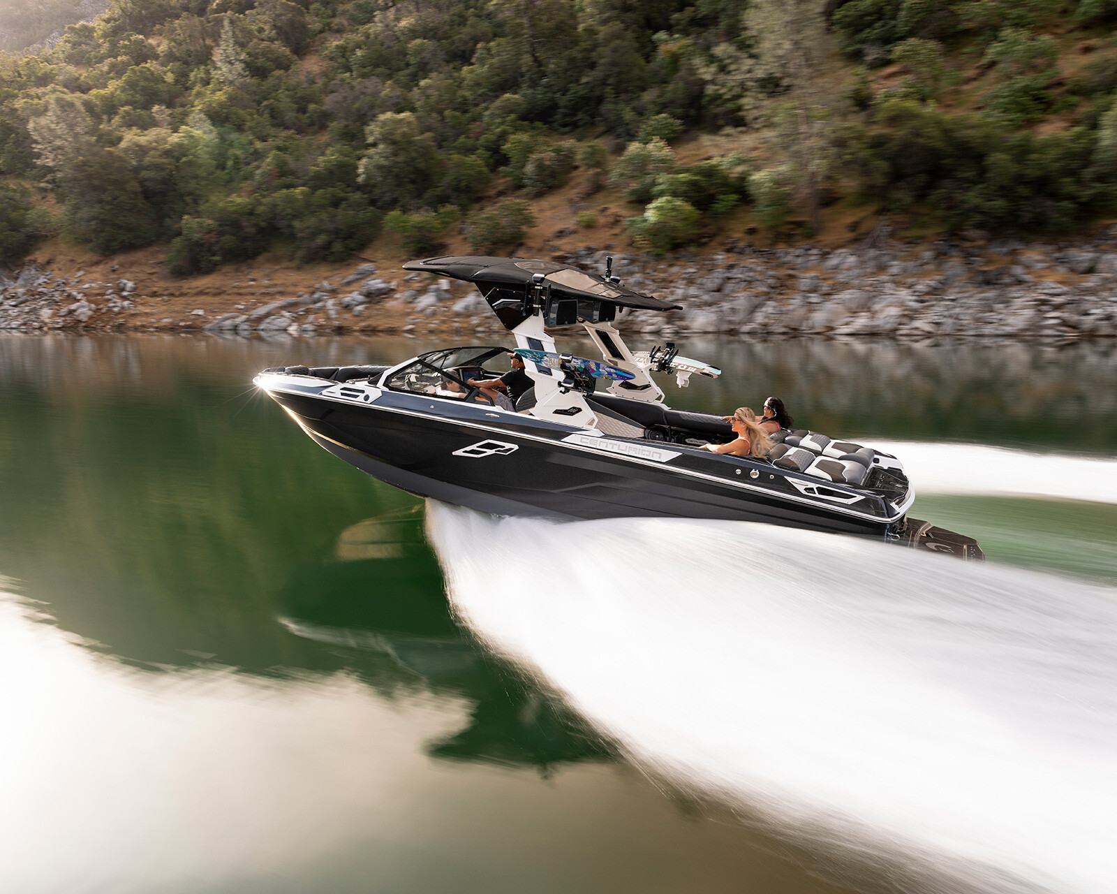 A Centurion Ri265 Wake Boat with several people on board speeds across a calm lake, creating a large wake; forested hills are visible in the background.