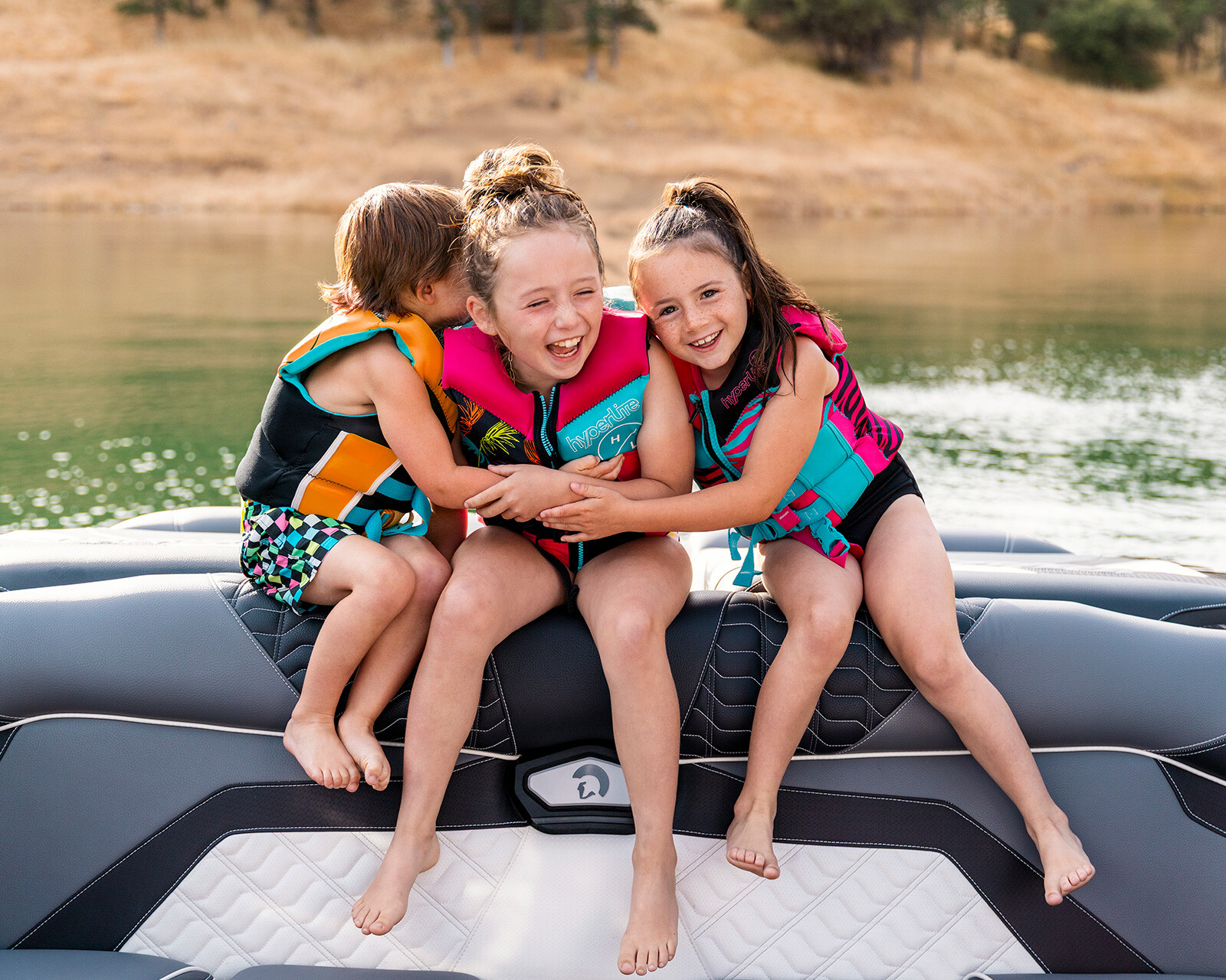 Three children wearing life jackets sit on a Centurion Ri265 boat seat by the water, smiling and hugging each other with a grassy shoreline in the background.