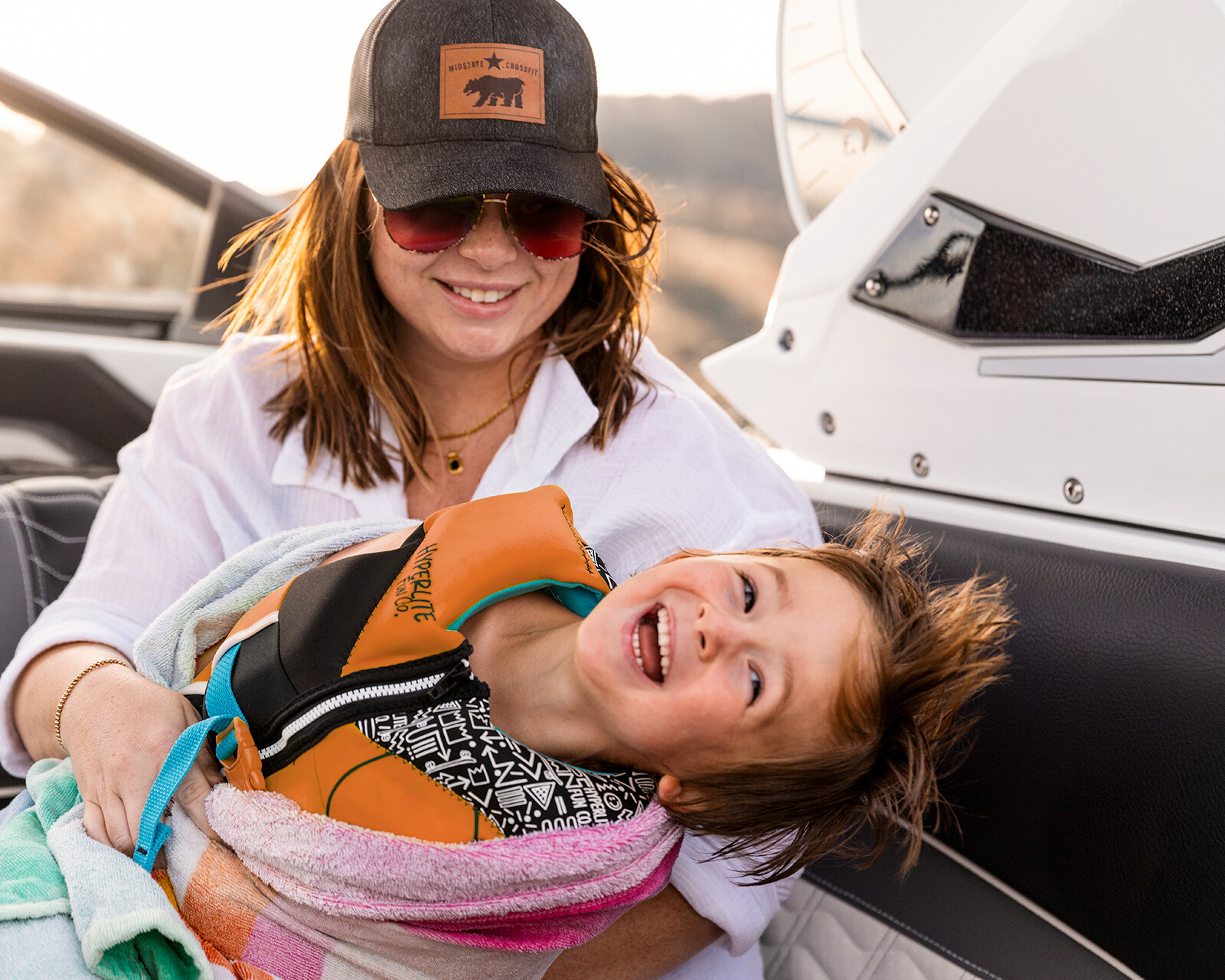 A woman in sunglasses and a hat holds a smiling child wrapped in a towel and wearing a life jacket on a Centurion Ri265 wake boat.