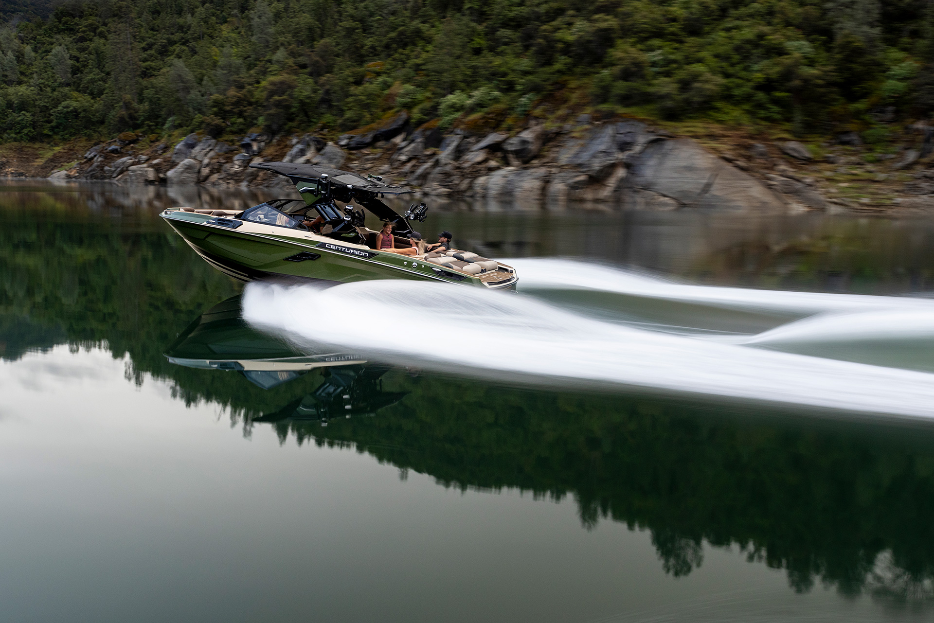 A Centurion Ri245 powerfully glides across a calm lake, leaving a trail of white wake behind, with forested hills and rocks in the background.