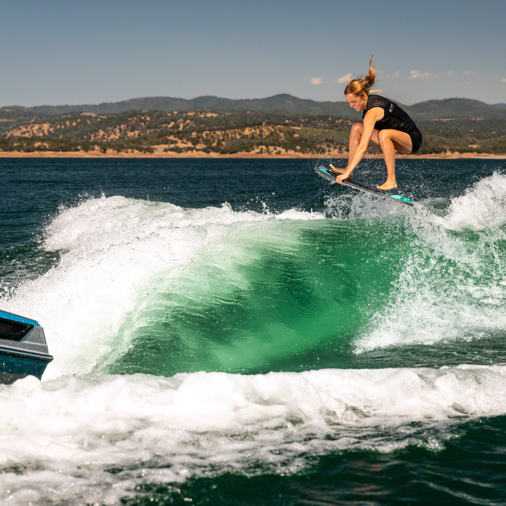 A person on a wakesurf board rides a wave behind a boat on a lake with distant tree-covered hills under a clear sky.