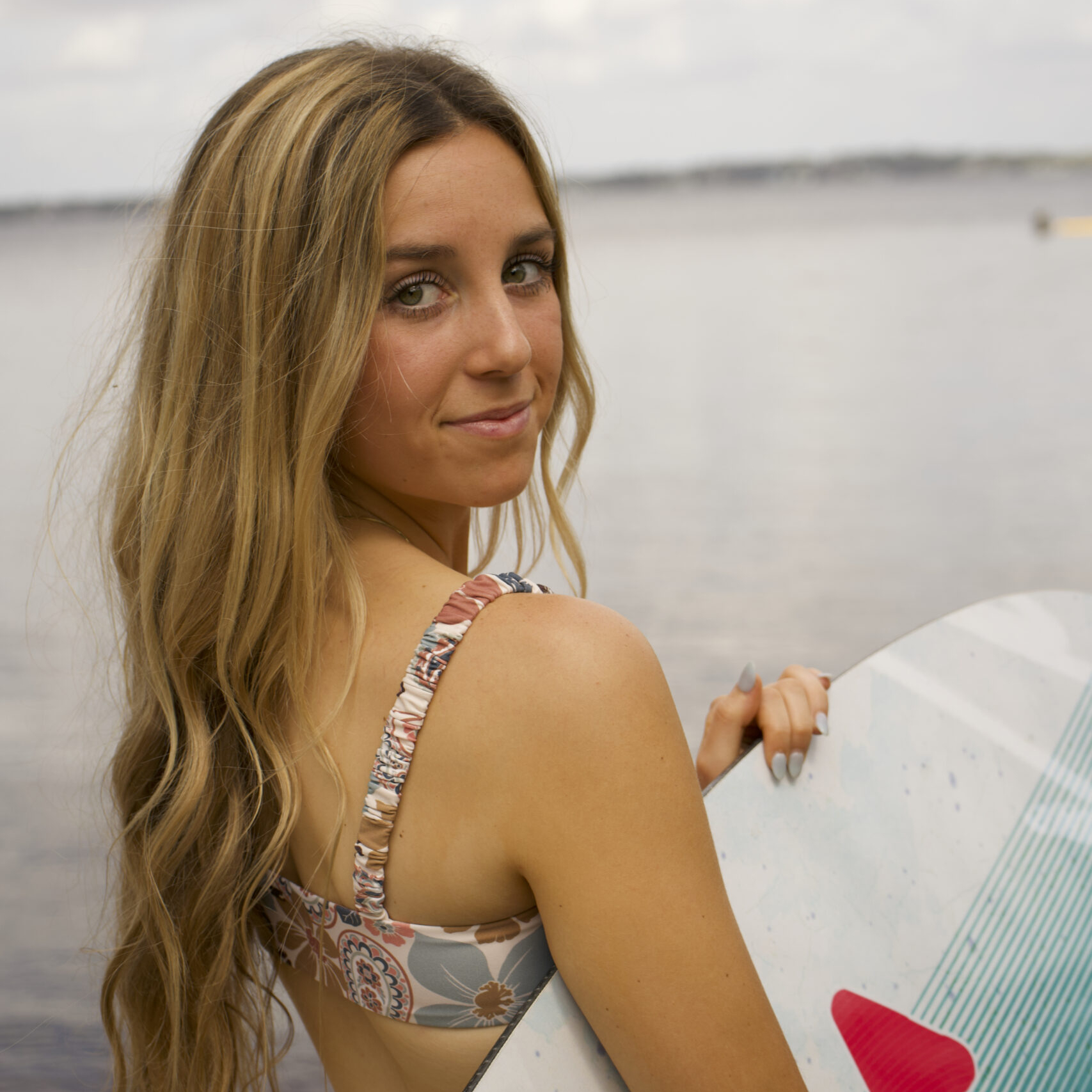 A young woman with long, wavy blonde hair stands by the water holding a surfboard, looking over her shoulder toward the camera.