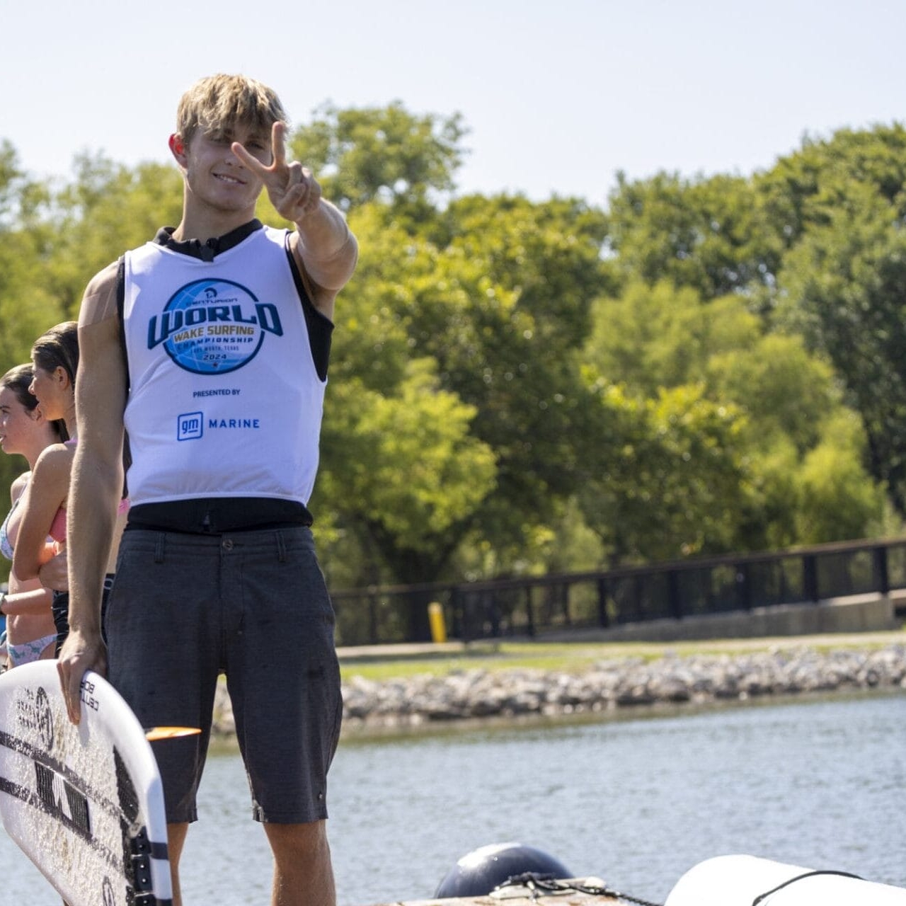 A person in a tank top holds up a peace sign beside a group of people near a waterfront.