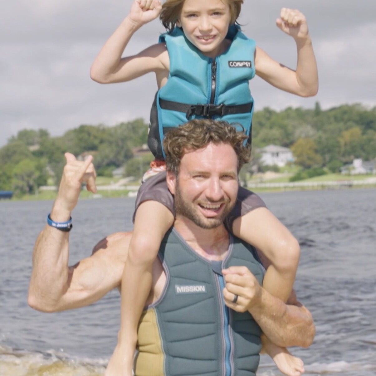An adult wearing a life vest carries a child on their shoulders at the edge of a body of water, with trees and houses visible in the background.