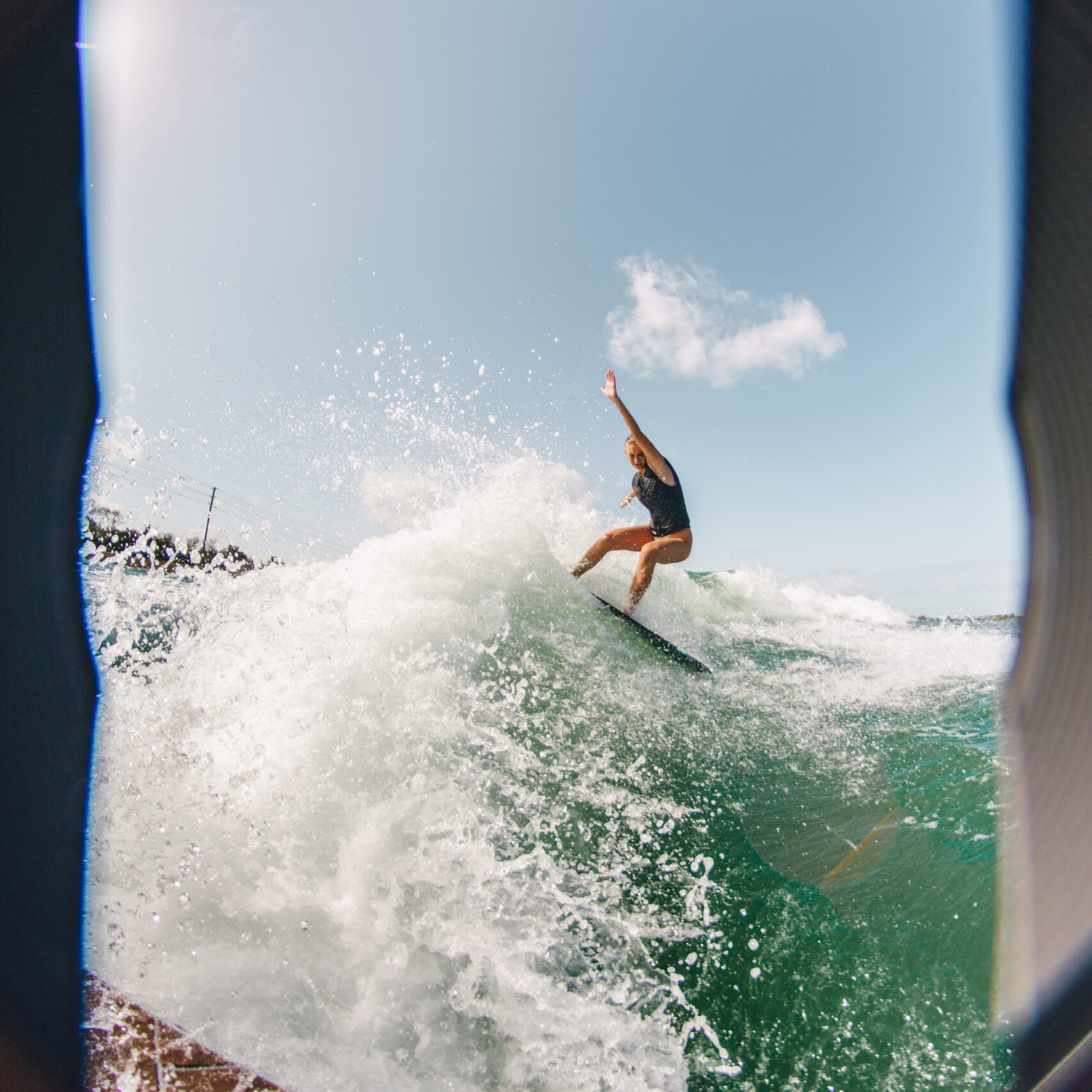 Ava Stewart rides a wave on a surfboard, captured from a low angle with water splashing and a bright blue sky in the background.