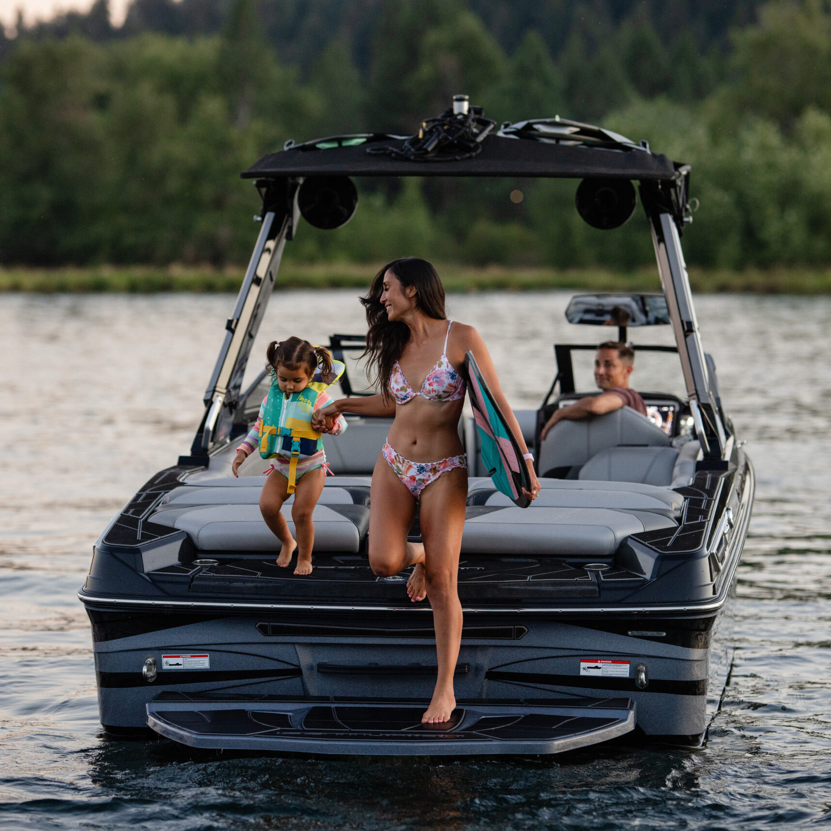 A woman holding a surfboard and a young child walk off the back of a boat onto the water, while another person sits inside the boat on a lake surrounded by trees.