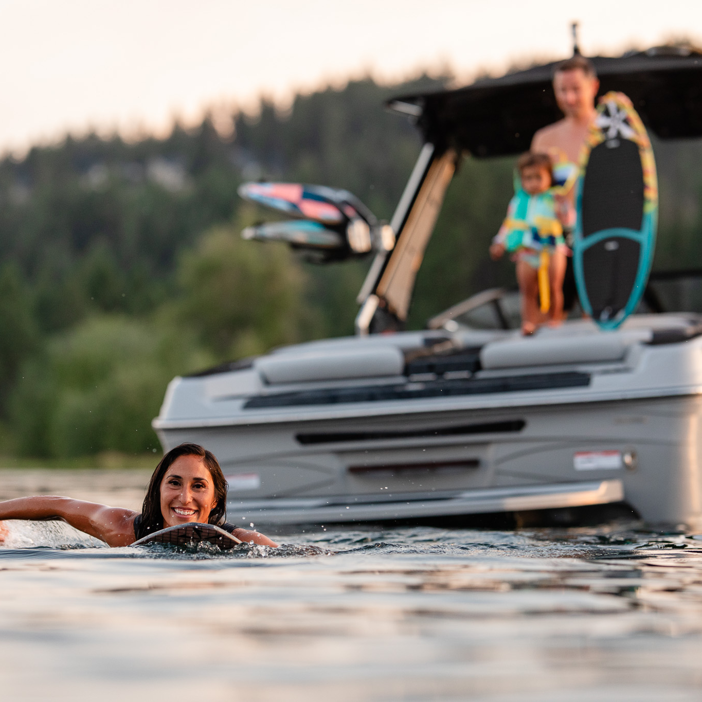 A woman swims in a lake near a boat, where an adult and a child holding a wakeboard stand on the rear deck. Trees and hills are visible in the background.