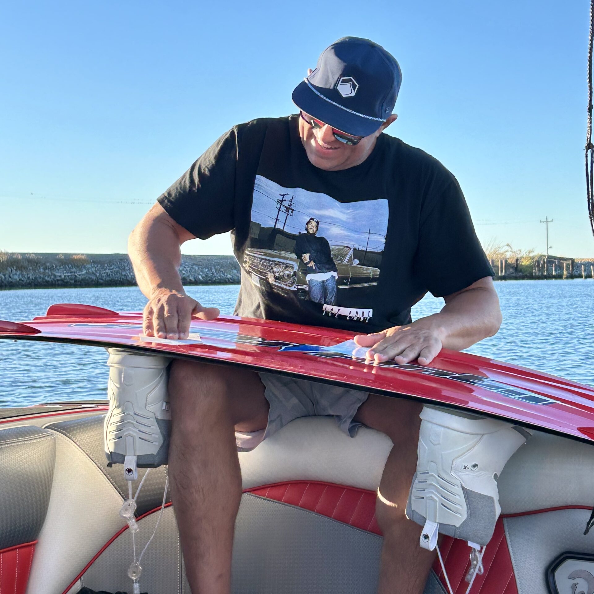 A man in a black T-shirt and hat sits on a boat seat, waxing a red wakeboard with a lake and blue sky in the background.