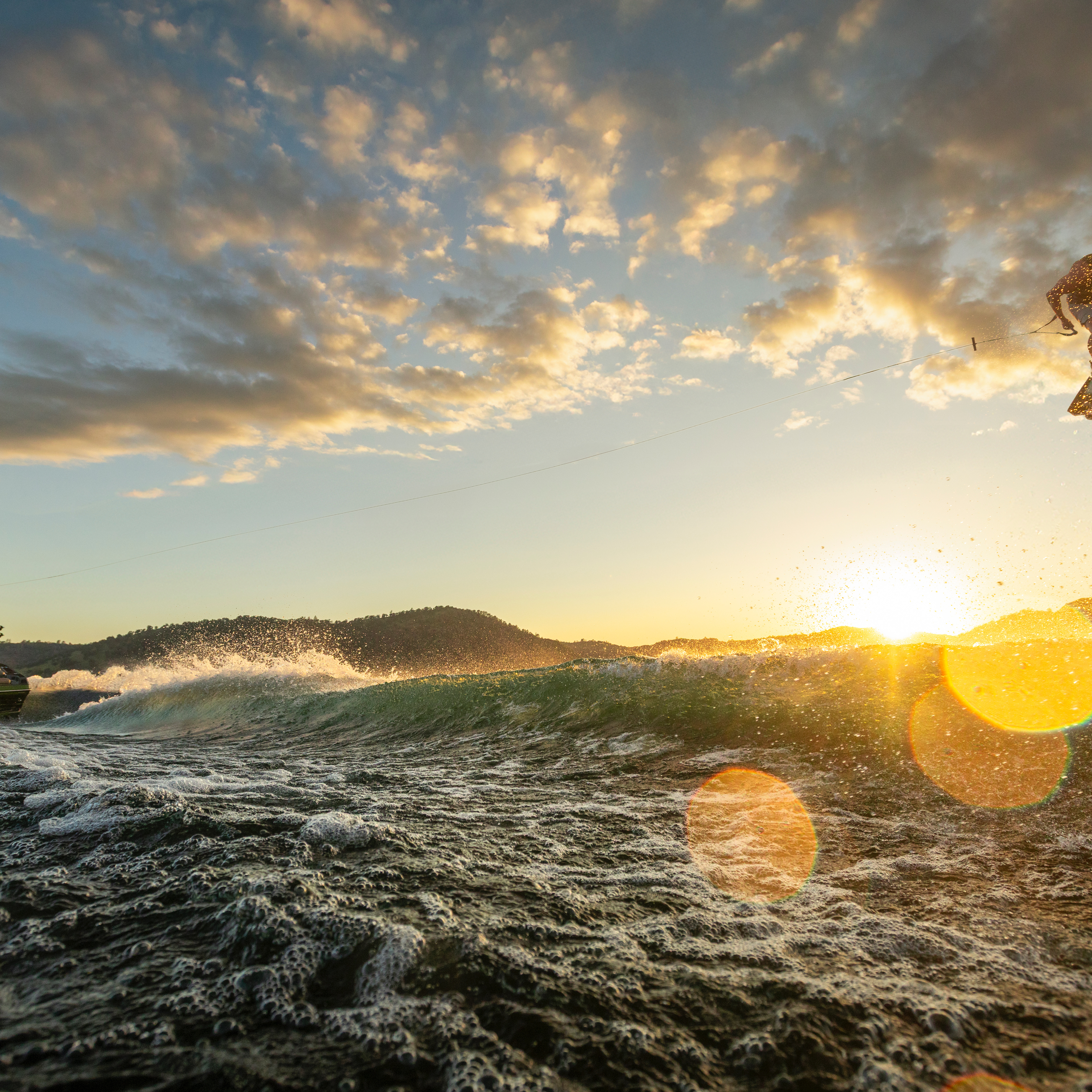 A person wakeboarding performs a jump behind a boat on a lake at sunset, with water spray and lens flares visible.