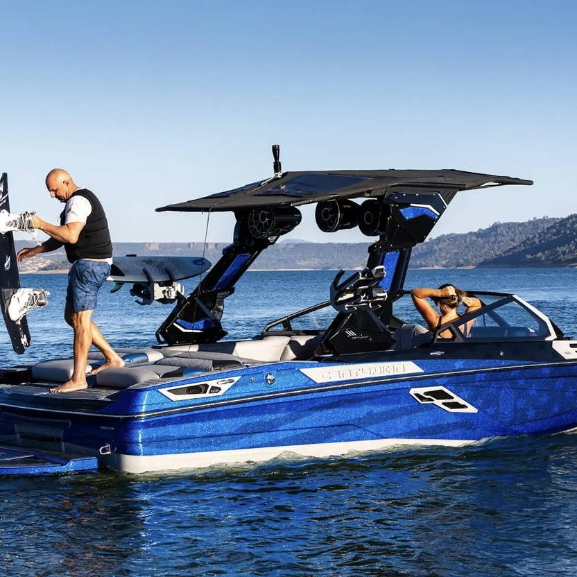 A man stands on the back of a blue motorboat holding a wakeboard, while another person sits near the helm on a sunny lake.