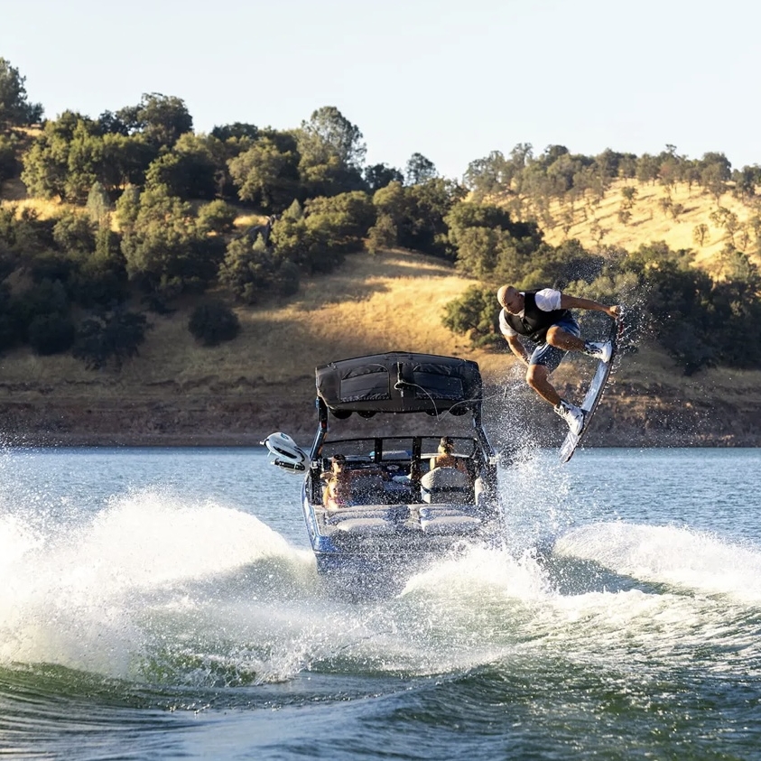 A person wakeboarding performs a jump behind a boat on a lake, with trees and hills visible in the background.
