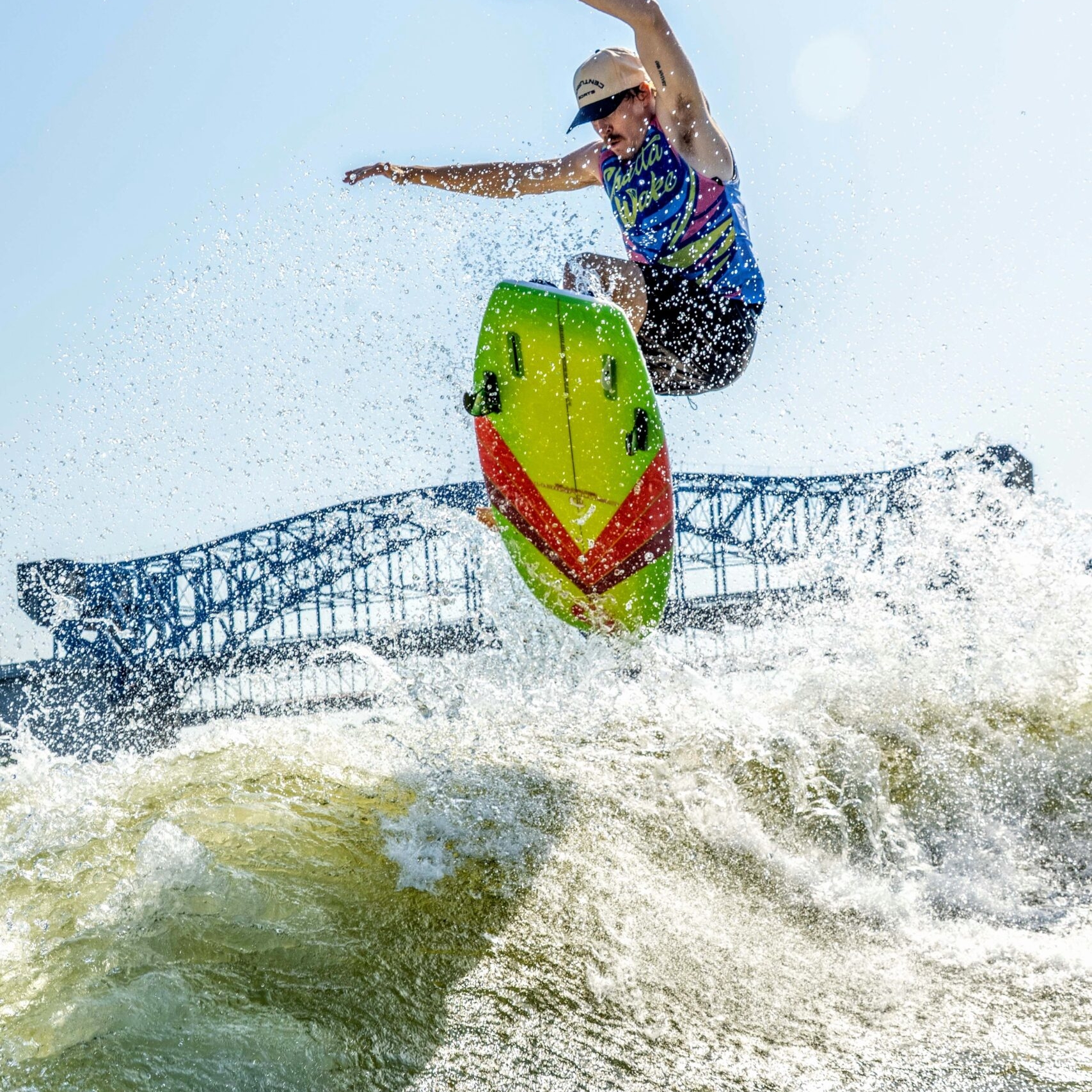 A person wearing a cap and life vest jumps a bright green and red jet ski over a wave, with a large bridge visible in the background.